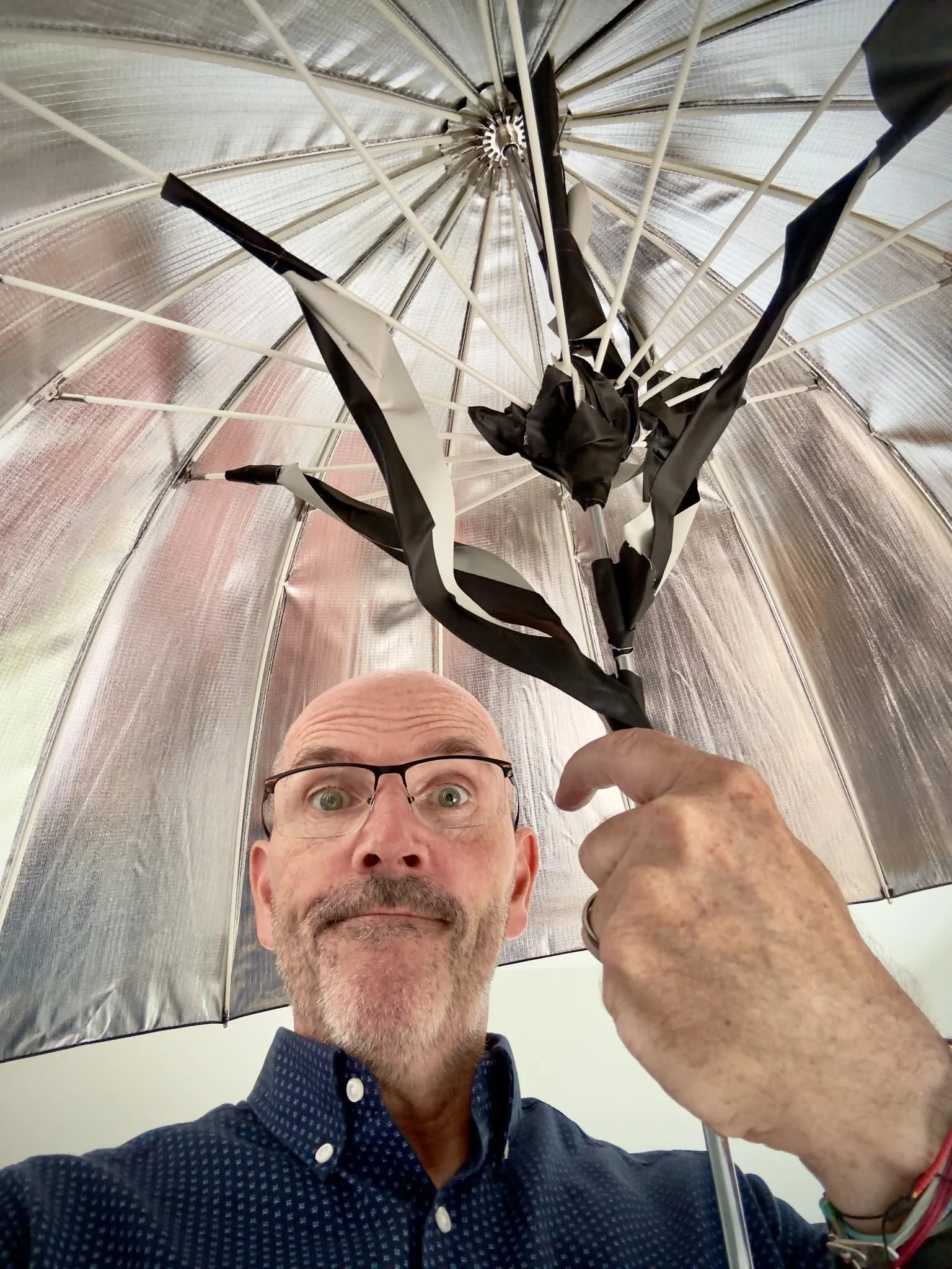 Photographer Mike Dick looking up wide-eyed at a silver photography umbrella with its snapped shaft held together with black gaffer tape, after a gust of wind blew over his lighting at a shoot in Barnet.