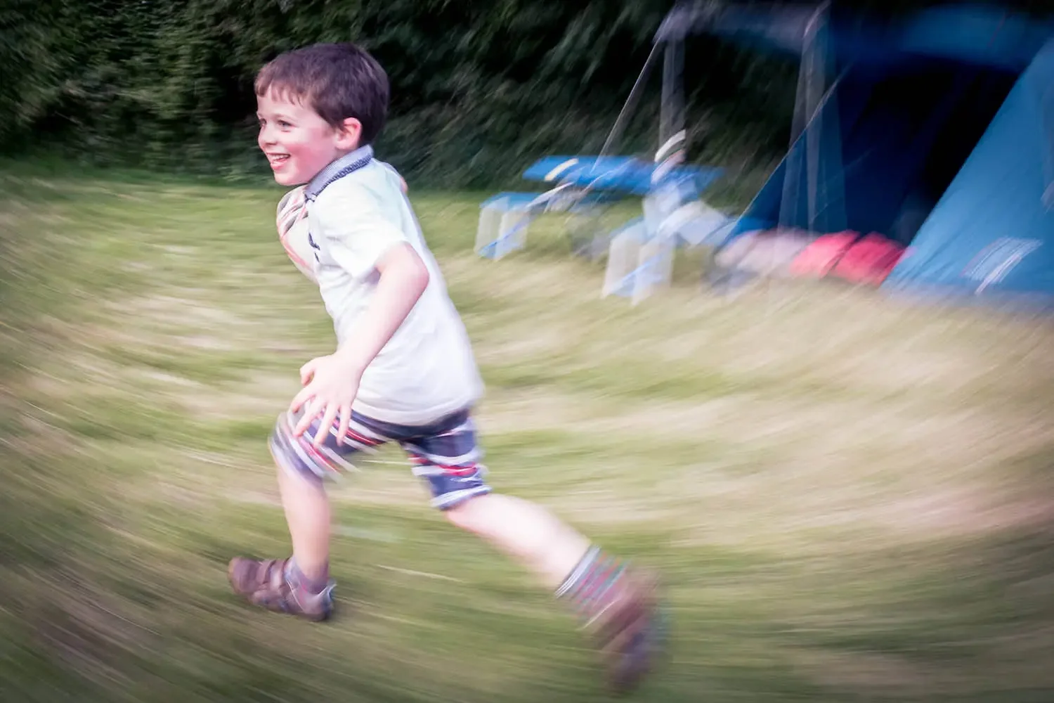 A young boy running outdoors across a grassy yard with a blue tent and outdoor furniture in the background, smiling and wearing a striped shirt and shorts.