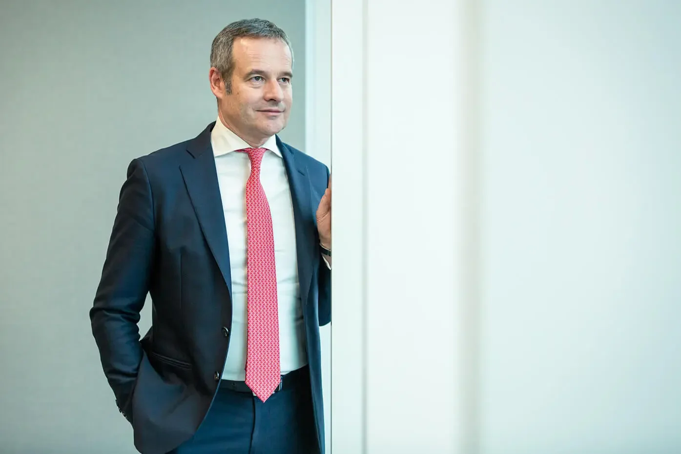A man in a business suit standing in an office, looking to the side, with a hand on a wall or doorframe.