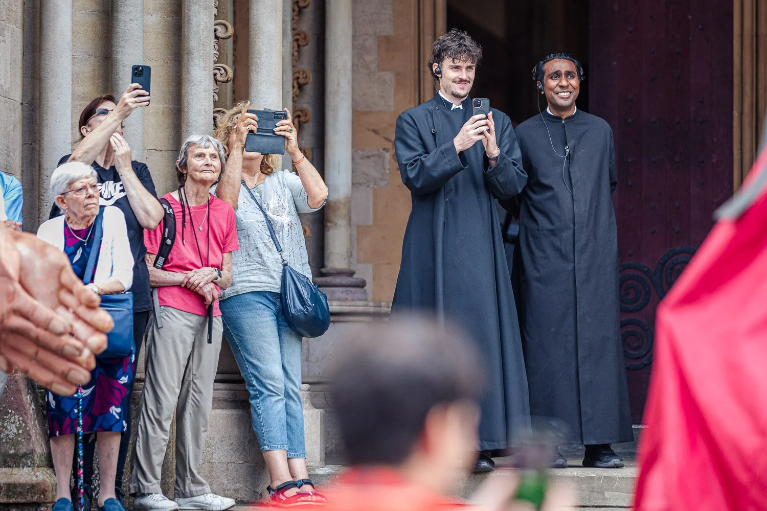 Crowd of spectators and two clergy in black cassocks photographing the Alban Day 2026 procession on the steps of St Albans Cathedral, Hertfordshire.
