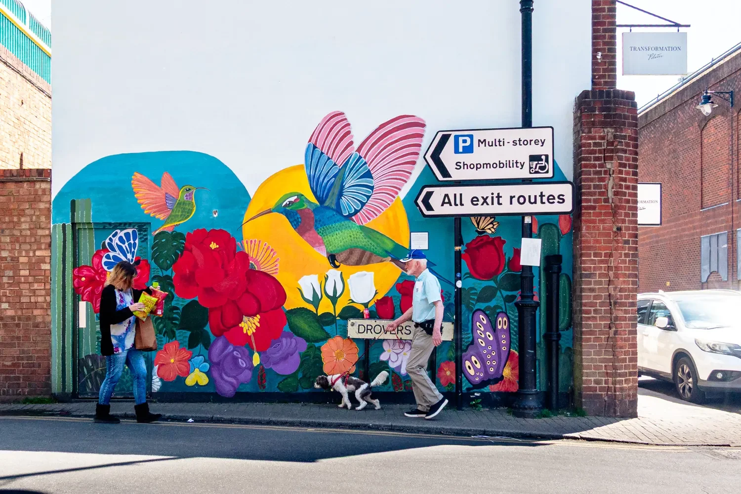 Colourful hummingbird and floral street mural on a building near Drovers in St Albans, with two pedestrians walking past in opposite directions