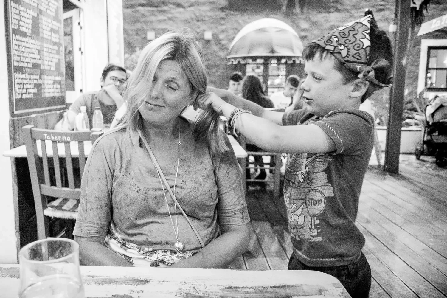 A boy with a party hat playfully tousling a woman's hair at a restaurant or cafe, with other people seated at tables in the background.