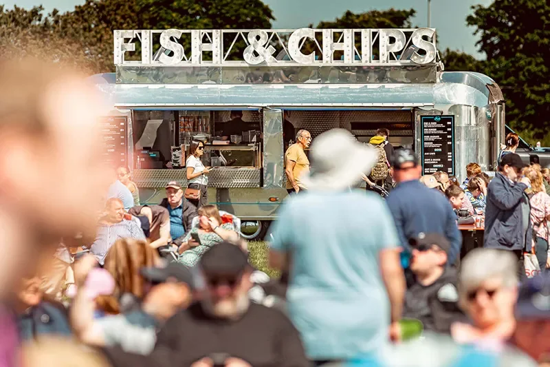 Crowded outdoor gathering with a fish and chips food truck under a sunny sky.