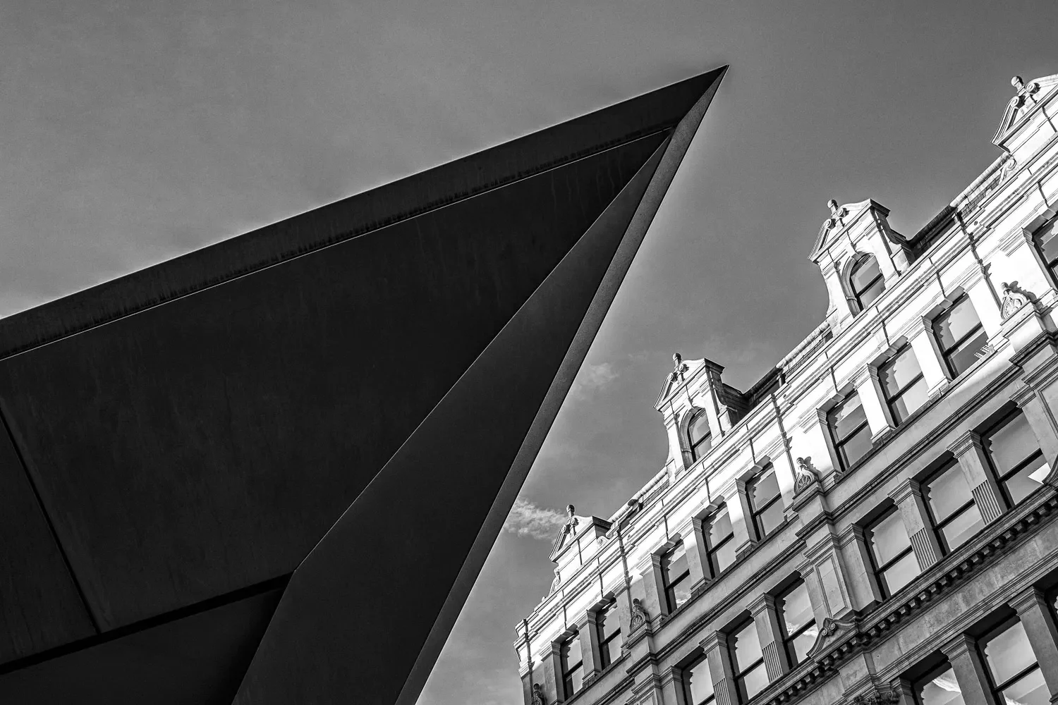 Sharp angular dark steel blade of a modern building cutting across an ornate white Victorian facade, shot from below, black and white