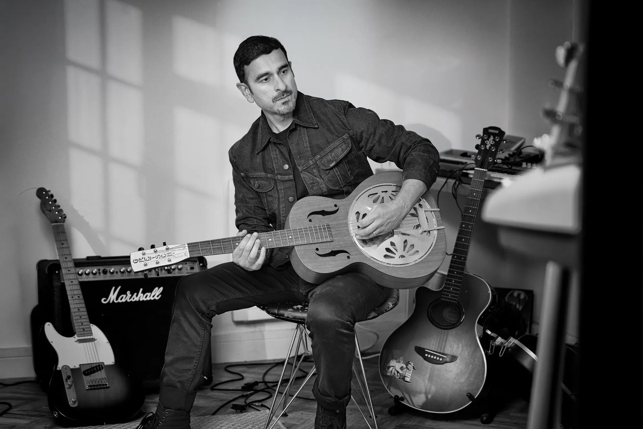 A man playing an acoustic resonator guitar in a music studio, surrounded by other guitars and music equipment.