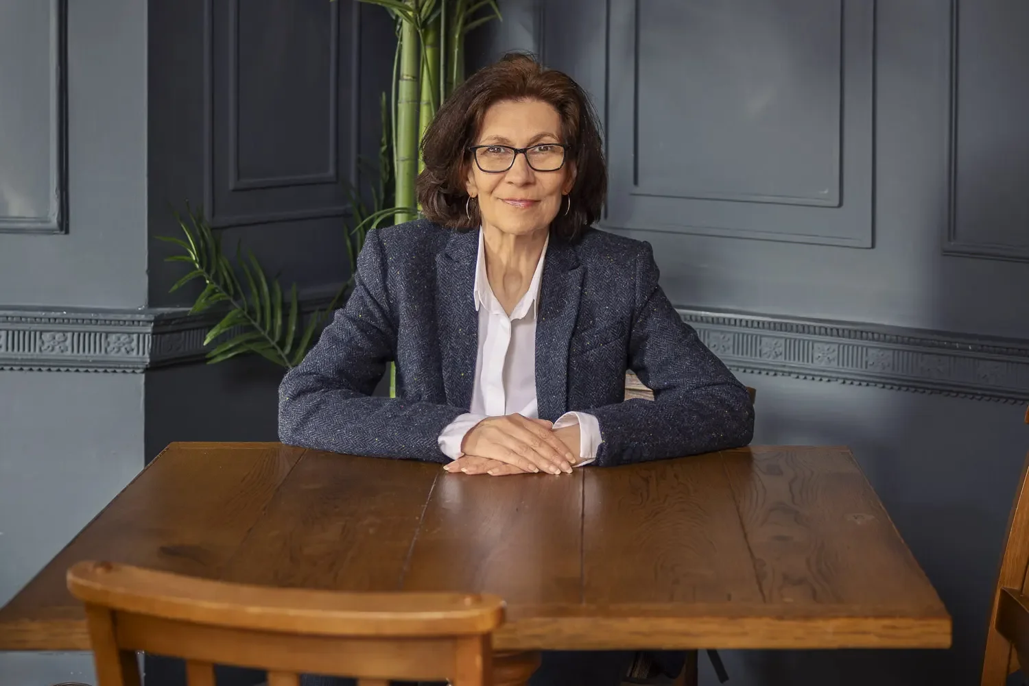 A middle-aged woman with short dark hair, glasses, and a white shirt sits at a wooden table, smiling at the camera in a room with dark blue wall paneling and green plants in the background.
