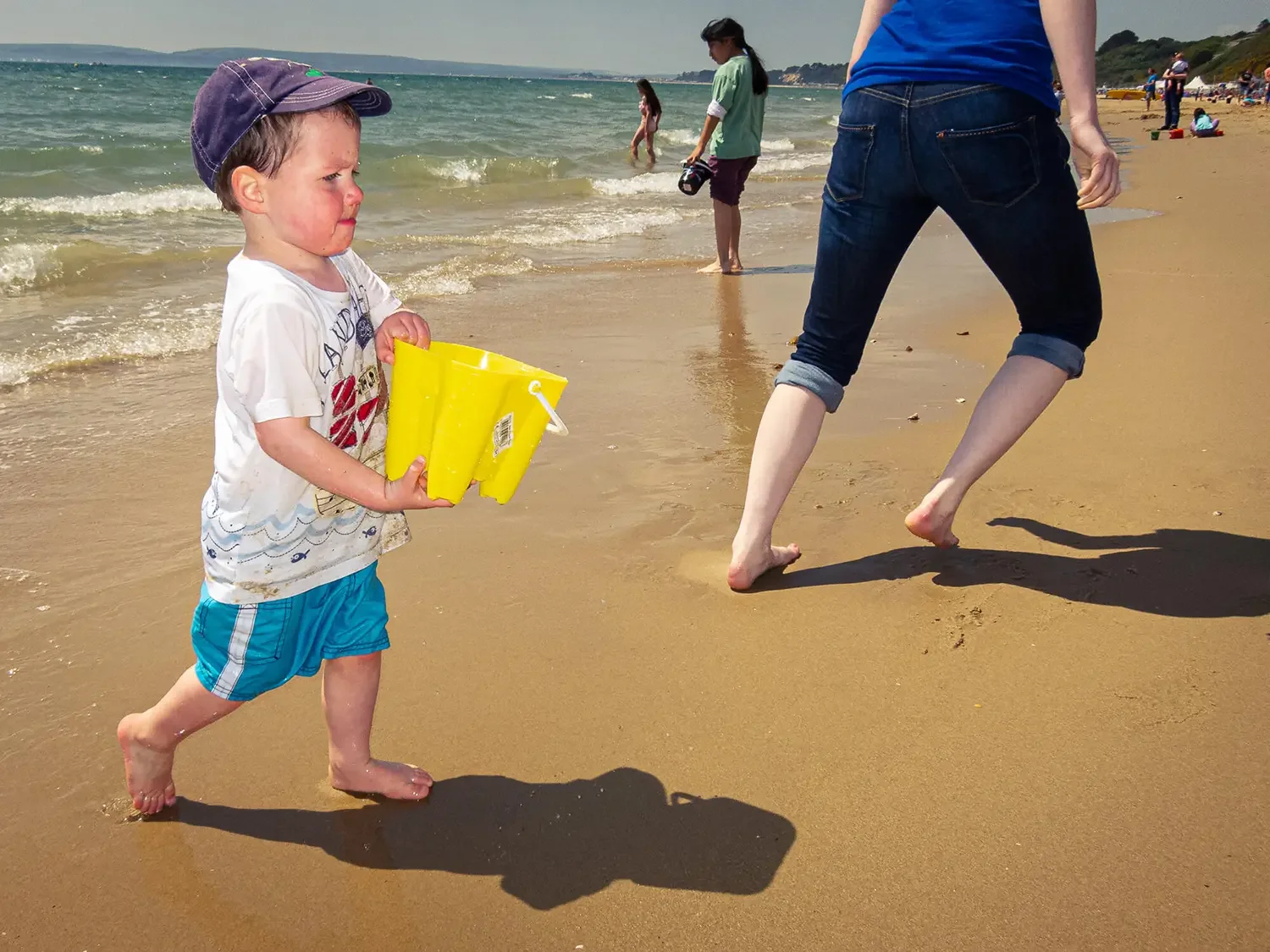 A young boy wearing a purple cap, a white T-shirt, and blue shorts, holding yellow buckets, walking along a sandy beach near the water. An adult with rolled-up jeans and a blue shirt walks ahead. Other people are visible in the background at the beac