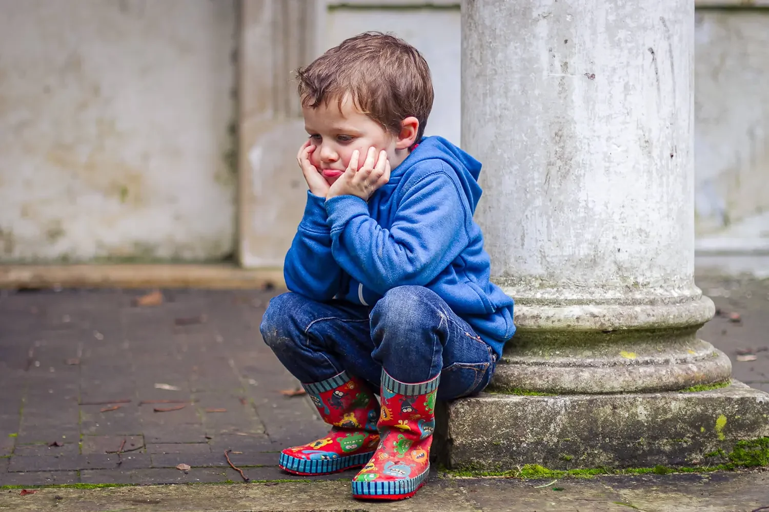 A young boy sitting on a stone step, looking upset, with hands on his face, wearing a blue hoodie and colorful rain boots, next to a large stone pillar.