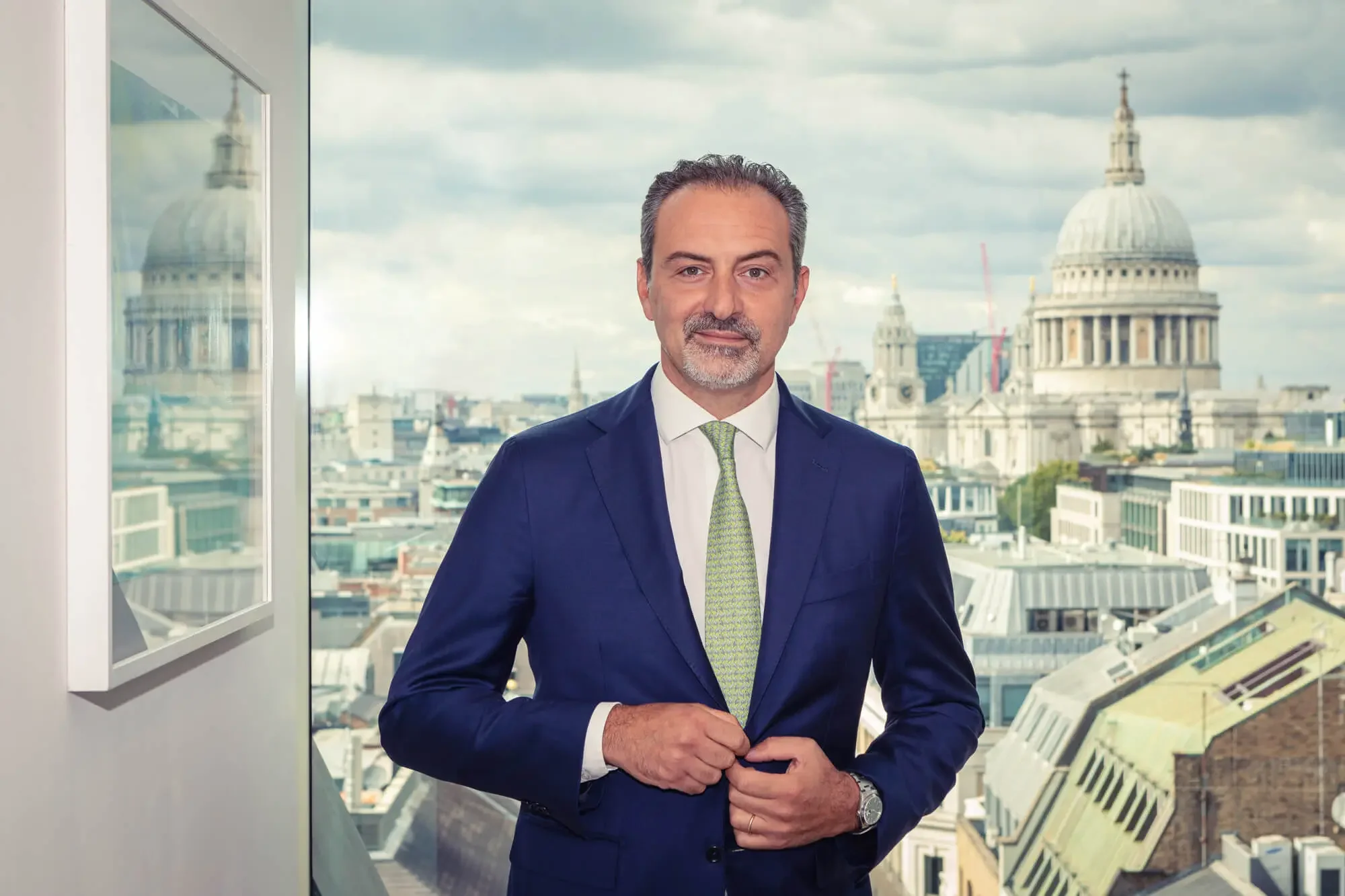 A man in a blue suit standing in front of a window with a city skyline and domed buildings, including St. Paul's Cathedral, in the background.