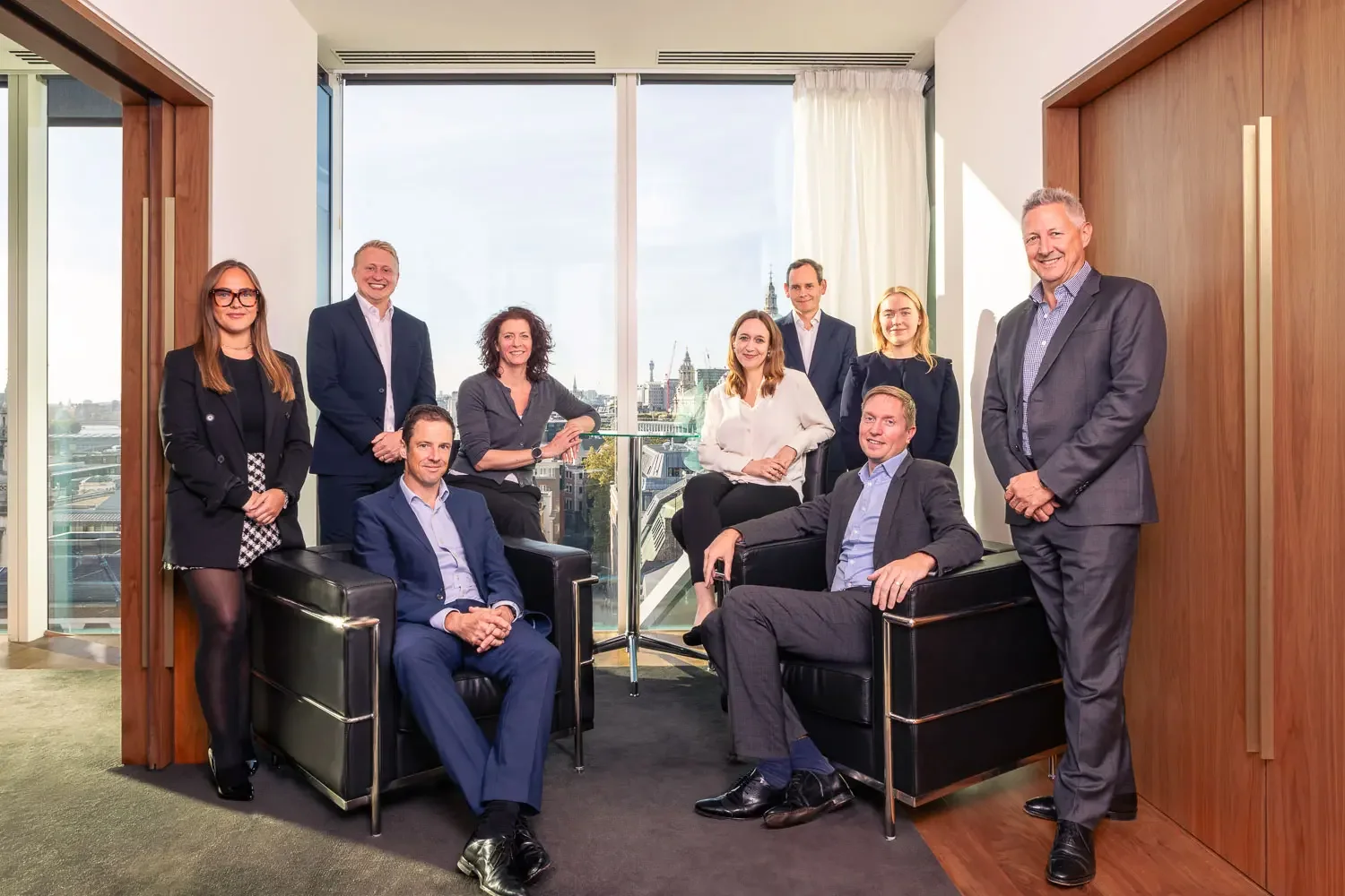 Group of nine professionals in a modern London office with large windows, St Pauls cathedral in the background city skyline view, two black armchairs, and a standing glass table.