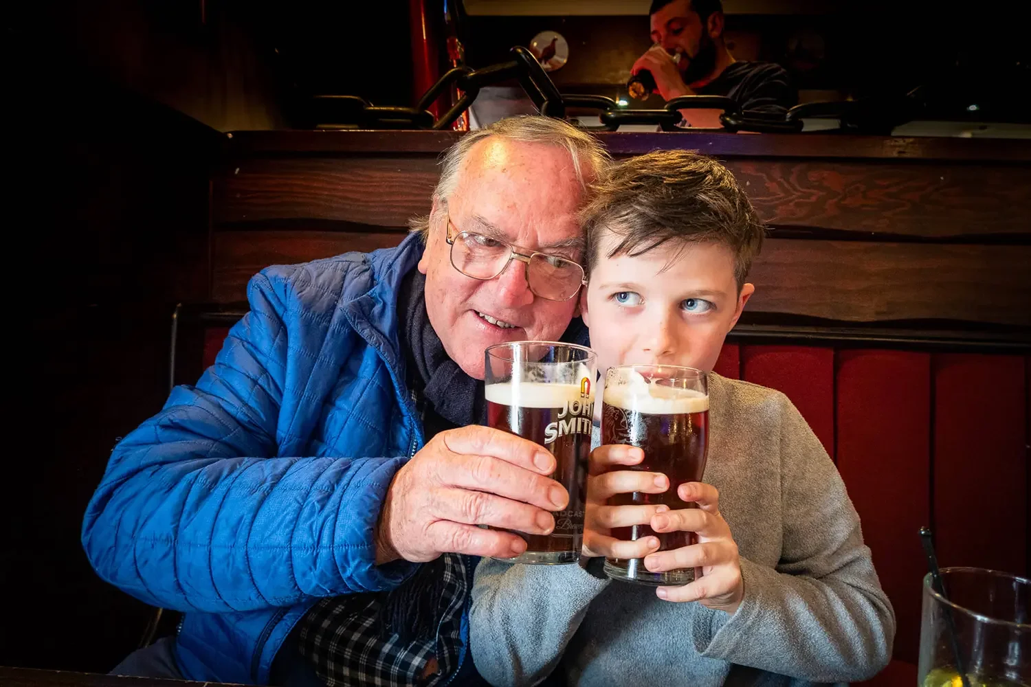An elderly man and a young boy, possibly a grandfather and grandson, are sitting at a table in a pub, holding glasses of dark beer. The man is smiling and wearing glasses and a blue jacket, while the boy appears to be looking sideways, dressed in a b