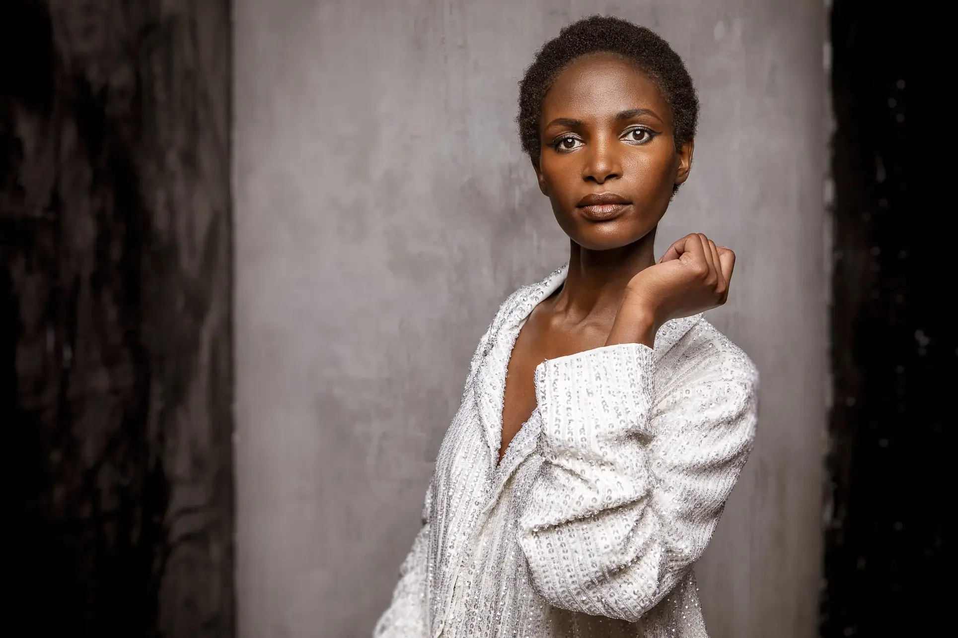 Portrait of a young Black woman with short natural hair, wearing a white textured jacket, standing against a neutral background, looking directly at the camera with a confident expression.