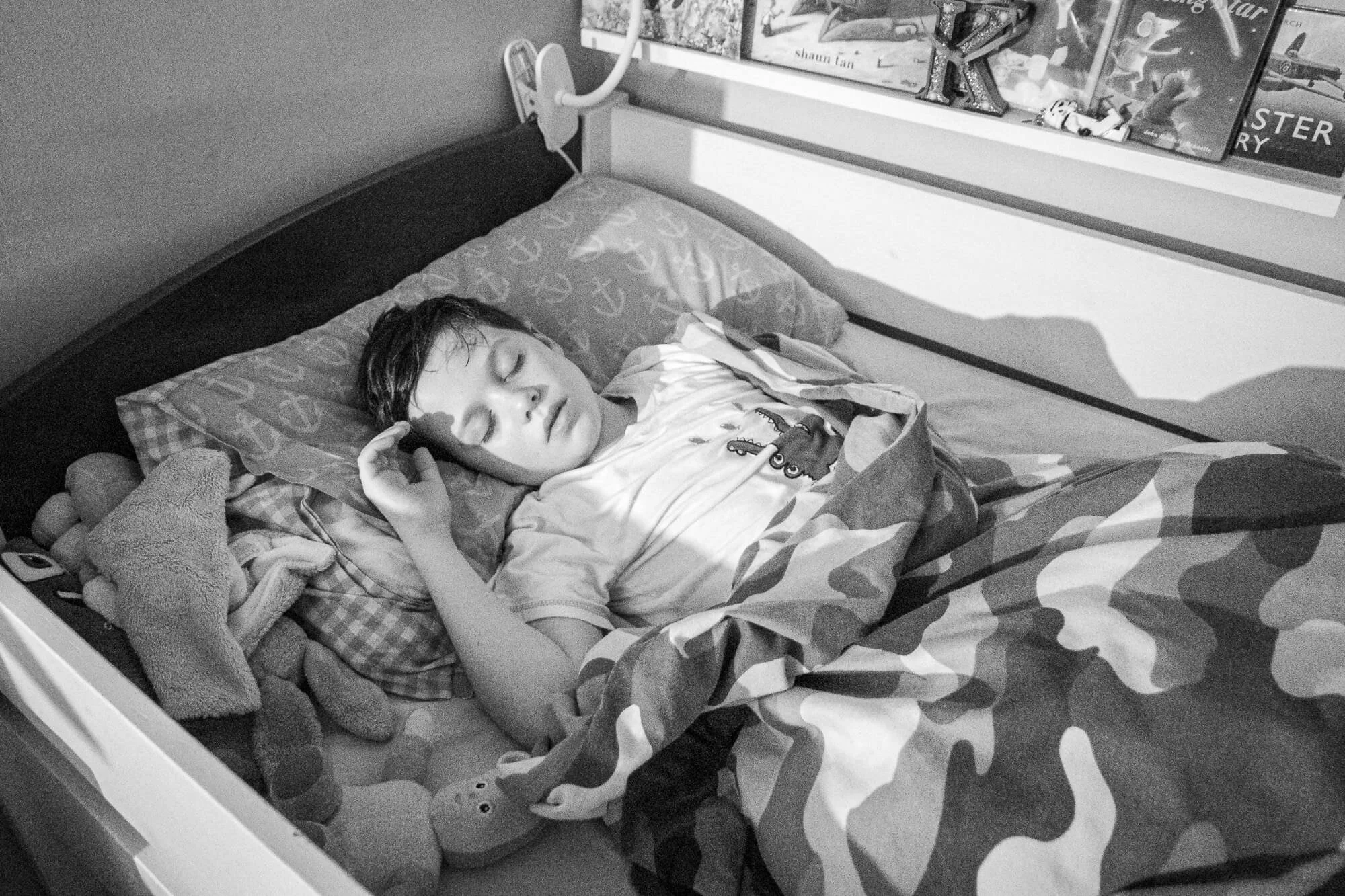 A young boy sleeping in a bed, holding a phone to his ear, with a stuffed animal beside him and books on the shelf above, in black and white.