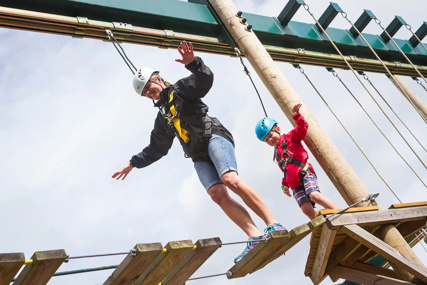 A woman and a young boy are on a wooden ropes course, wearing safety helmets and harnesses. The woman is balancing on a wooden plank suspended in midair, smiling and waving, while the boy is behind her, also balancing and waving. The background shows