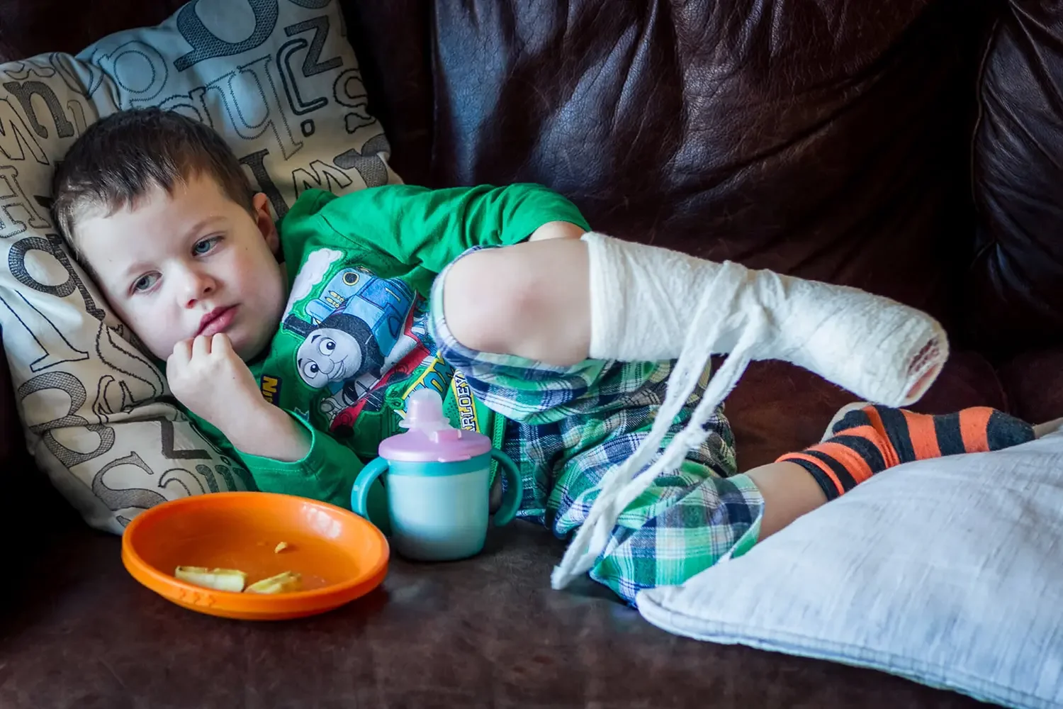 A young boy with blue eyes and short brown hair lying on a sofa, resting his head on a patterned pillow. He is wearing a green Thomas the Tank Engine shirt and plaid shorts, with one leg elevated and wrapped in a white bandage. In front of him is a l