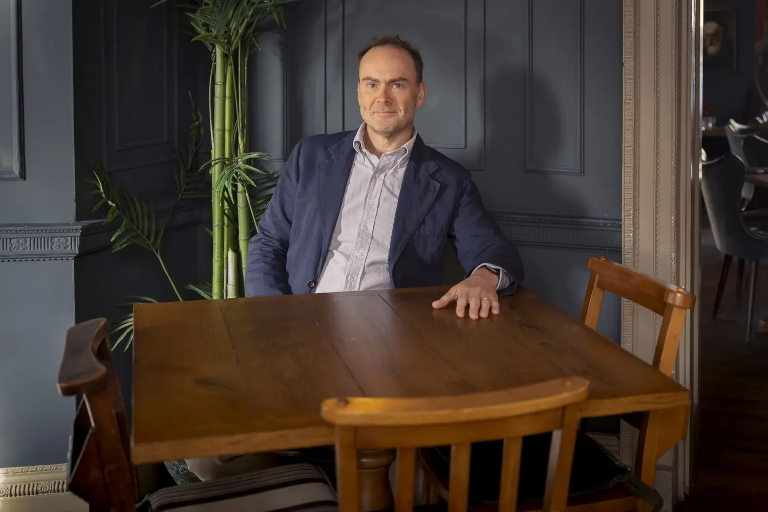 A man with short dark hair and a slight beard, wearing a light-colored shirt and a dark blazer, sitting at a wooden table in a room with dark blue walls and paneling. There is a large green plant behind him and a chair in front of the table.