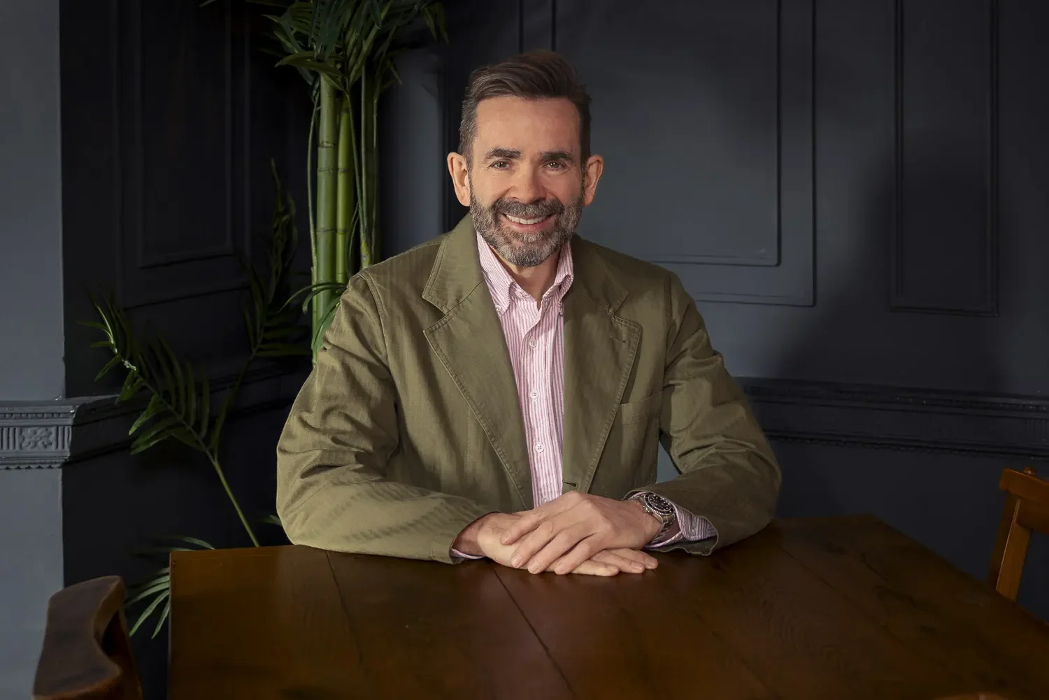 A man with a beard and short hair, wearing a light pink striped shirt and khaki jacket, sitting at a wooden table in a dark, stylish room with black paneled walls and a green plant in the background, smiling at the camera.
