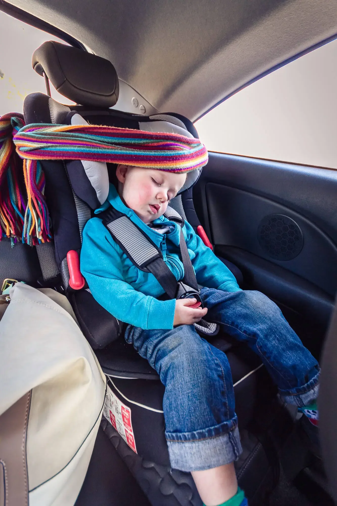 A young boy sleeping in a car seat inside a vehicle, wearing a blue jacket and jeans, with a colorful scarf on top of the headrest.
