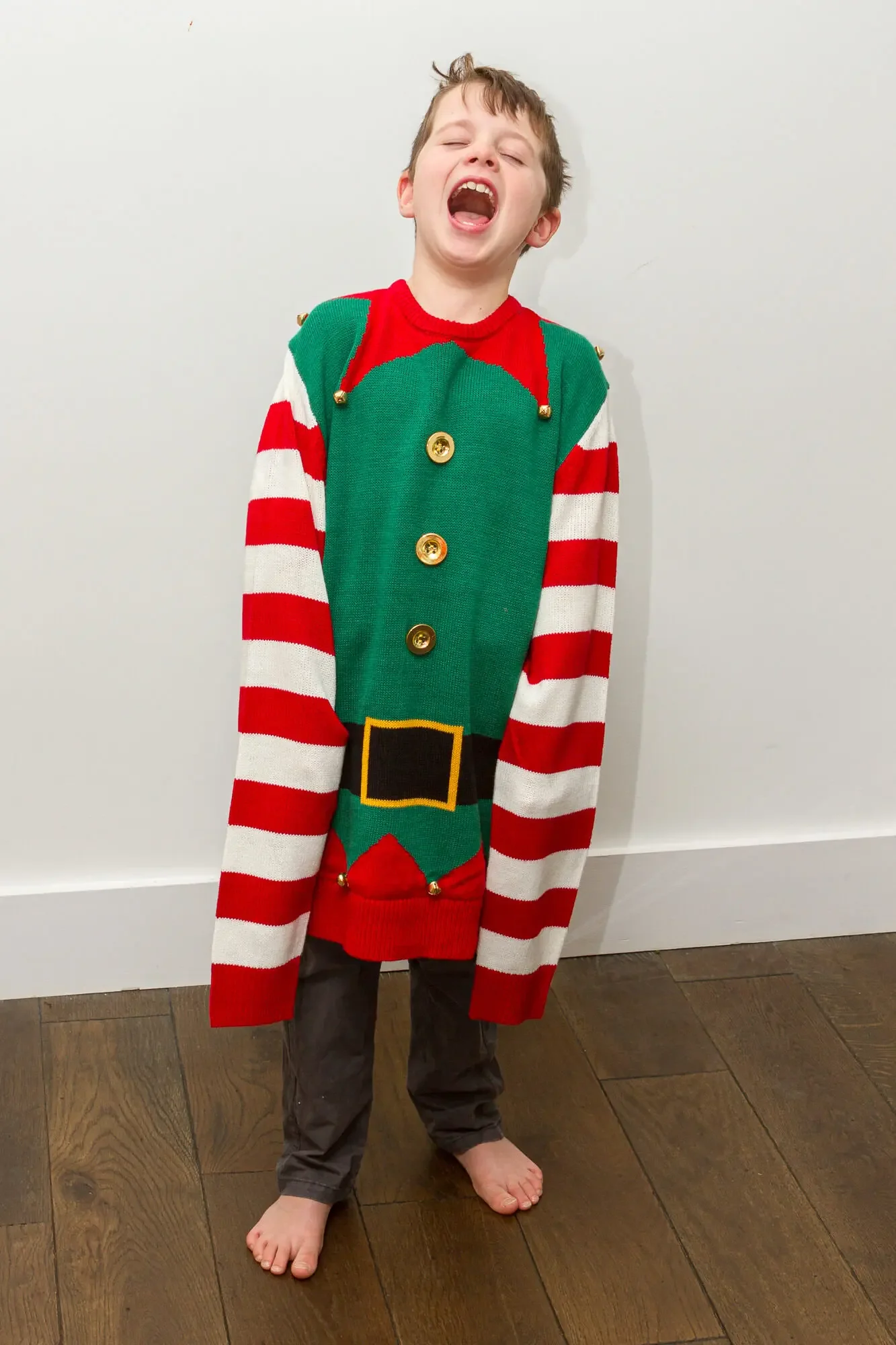 A young boy laughing and standing barefoot against a white wall, wearing a festive elf costume with red and white striped sleeves, green front with black belt, and gold bells.