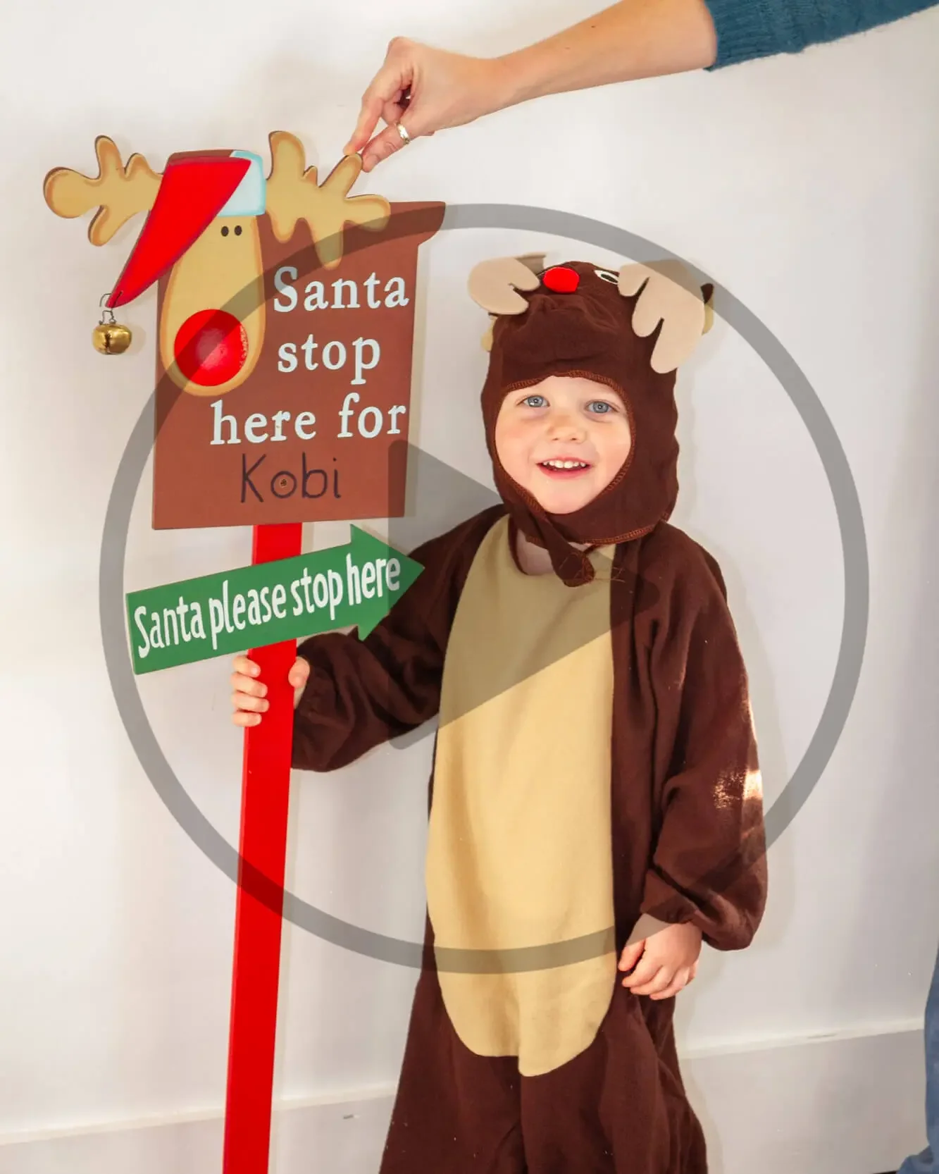 Young child dressed in a reindeer costume, standing next to a festive sign that says 'Santa stop here for Kobi' with an arrow pointing to himself, and a hand adjusting a Santa hat on a reindeer figure.