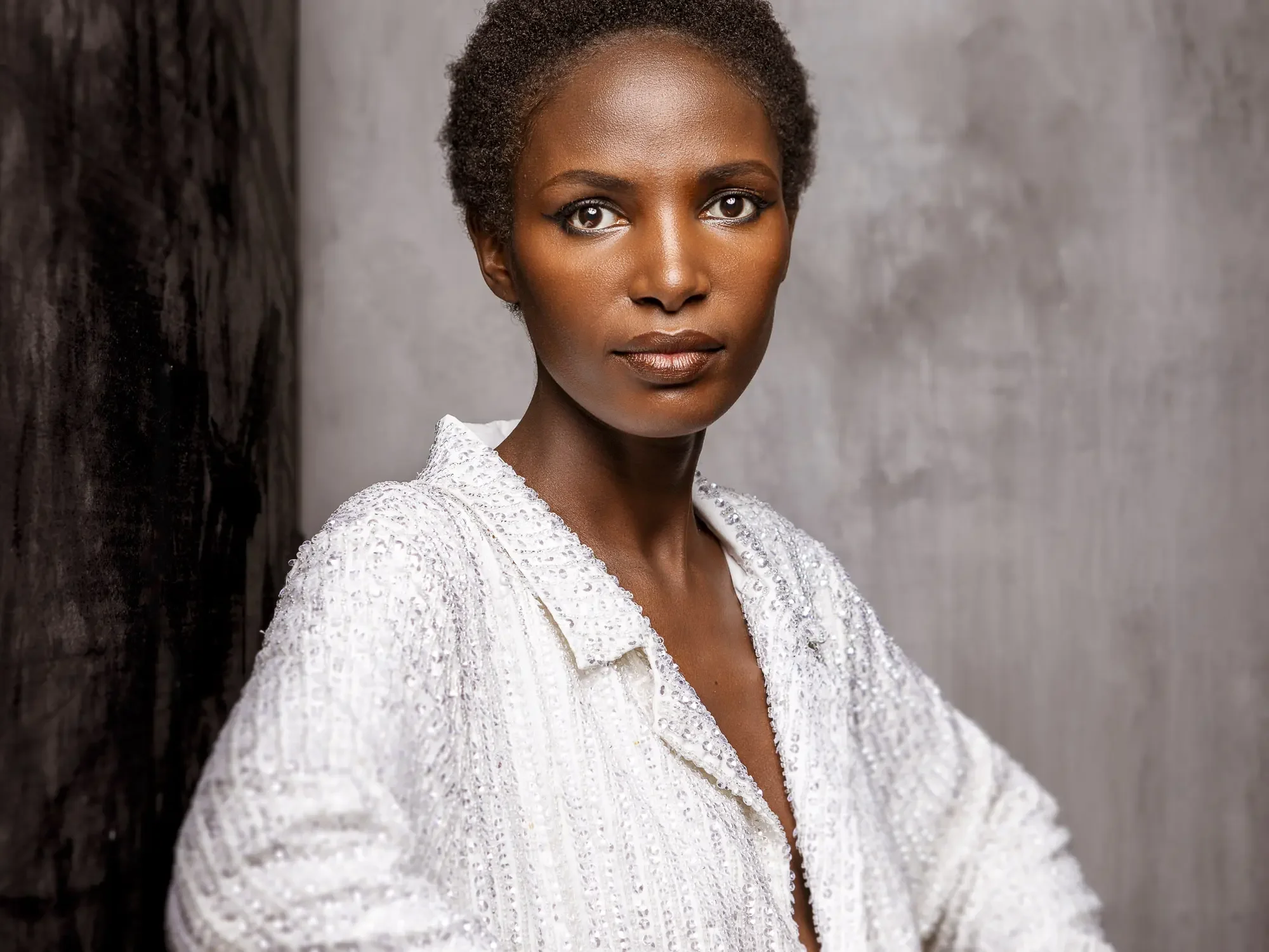 Portrait of a woman with short curly hair, wearing a white textured jacket, standing against a gray and dark background.
