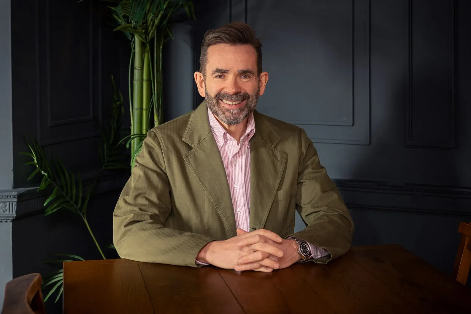 Male business professional in an olive blazer seated at a wooden table against a dark blue panelled wall — personal branding headshot photography by Mike Dick Photography, St Albans