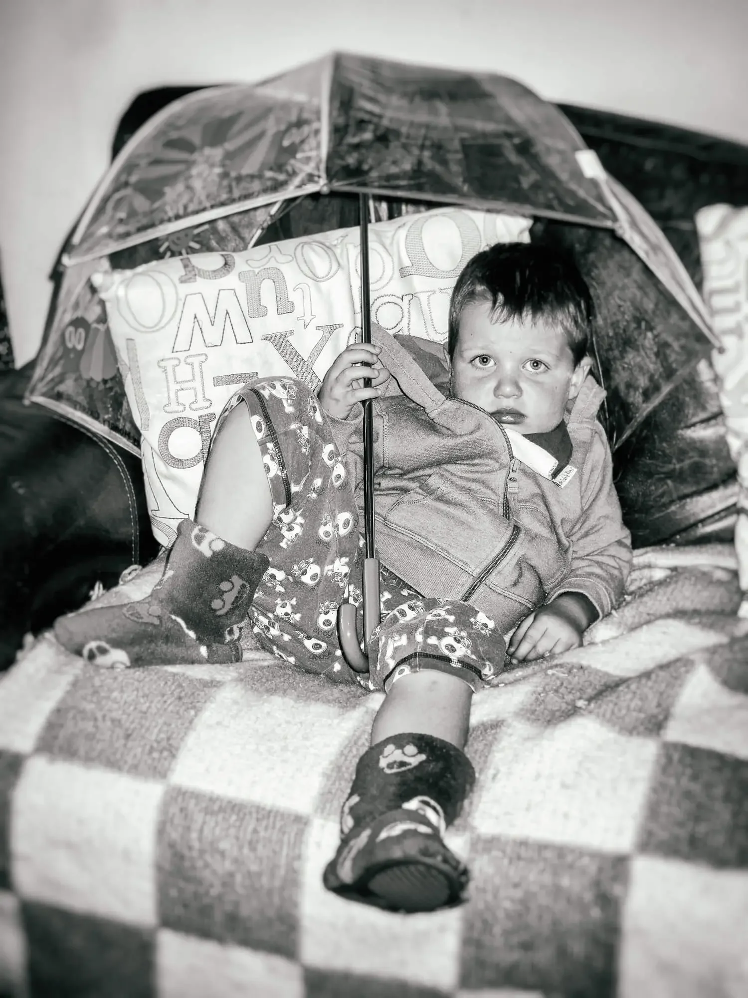 Black and white photo of a young boy sitting on a blanket, holding an open umbrella. The boy is wearing a jacket, patterned pajama pants, and socks with skull designs, looking directly at the camera.