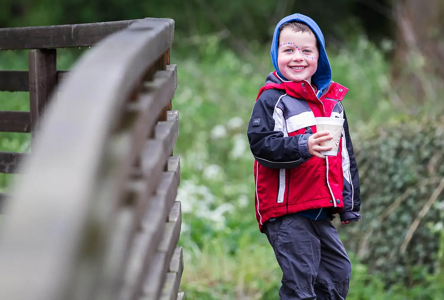 A smiling young boy in a colorful jacket and face paint holds a cup while standing outdoors near a wooden fence, with a green, leafy background.