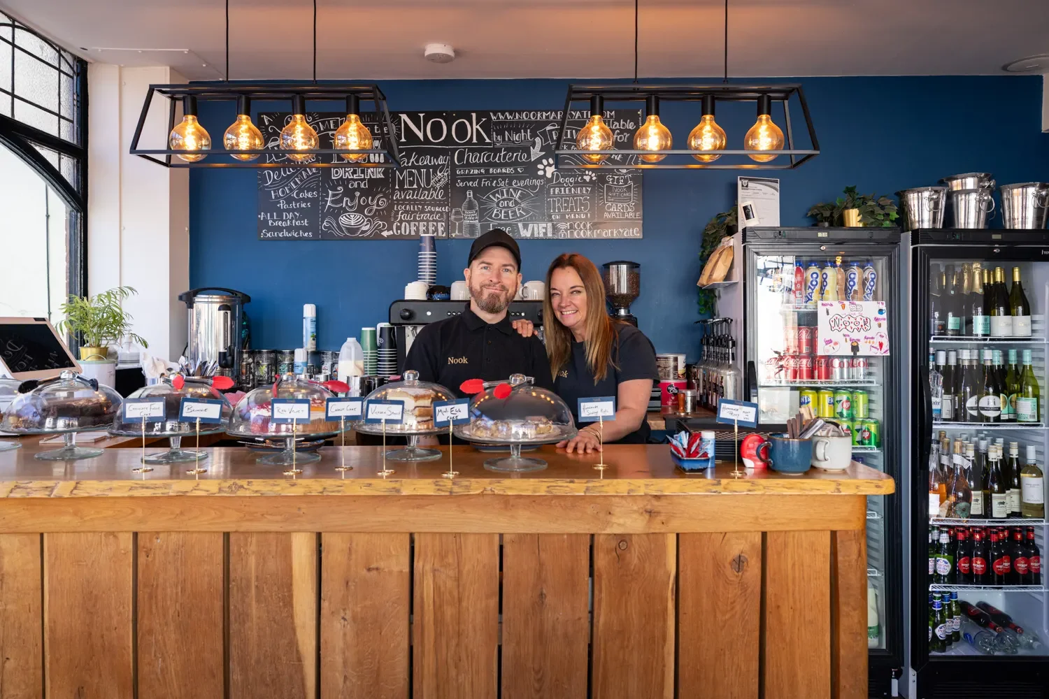 Wide interior shot of the Nook café in Markyate showing the full cake counter, Edison bulb lighting, chalkboard menus, and the two owners — brand photography by Mike Dick Photography