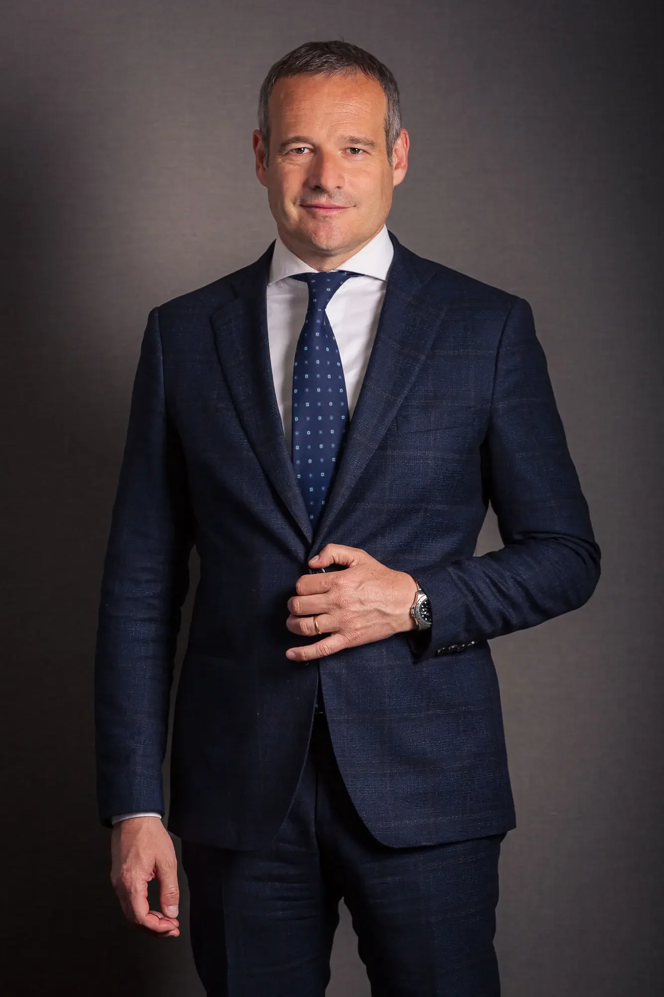 A man in a dark blue suit, white shirt, and patterned blue tie standing against a gray background. He is adjusting his suit jacket with his right hand and wears a watch on his left wrist.
