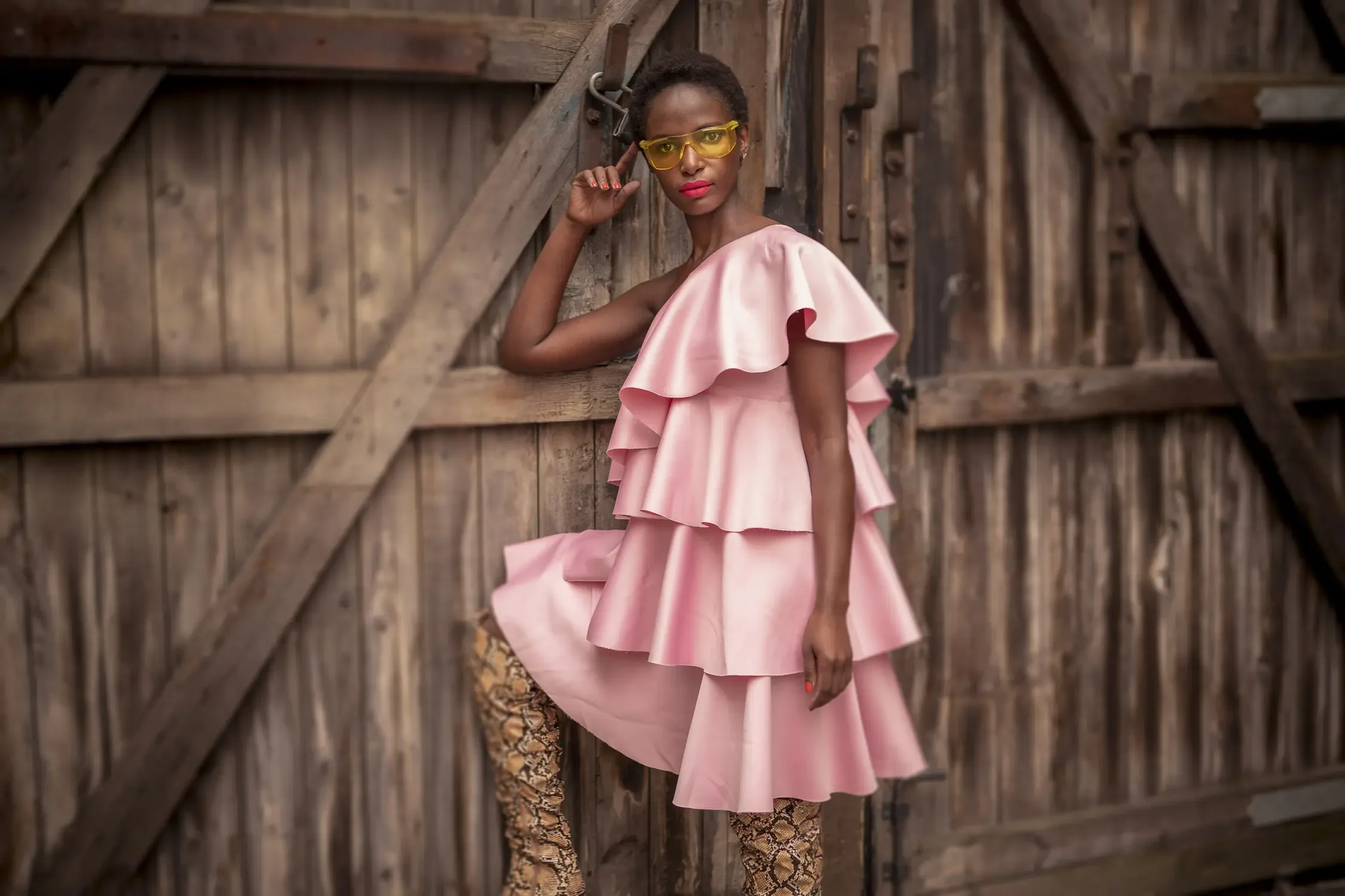 A woman wearing a pink ruffled dress, yellow sunglasses, and patterned tights standing against a wooden barn door.