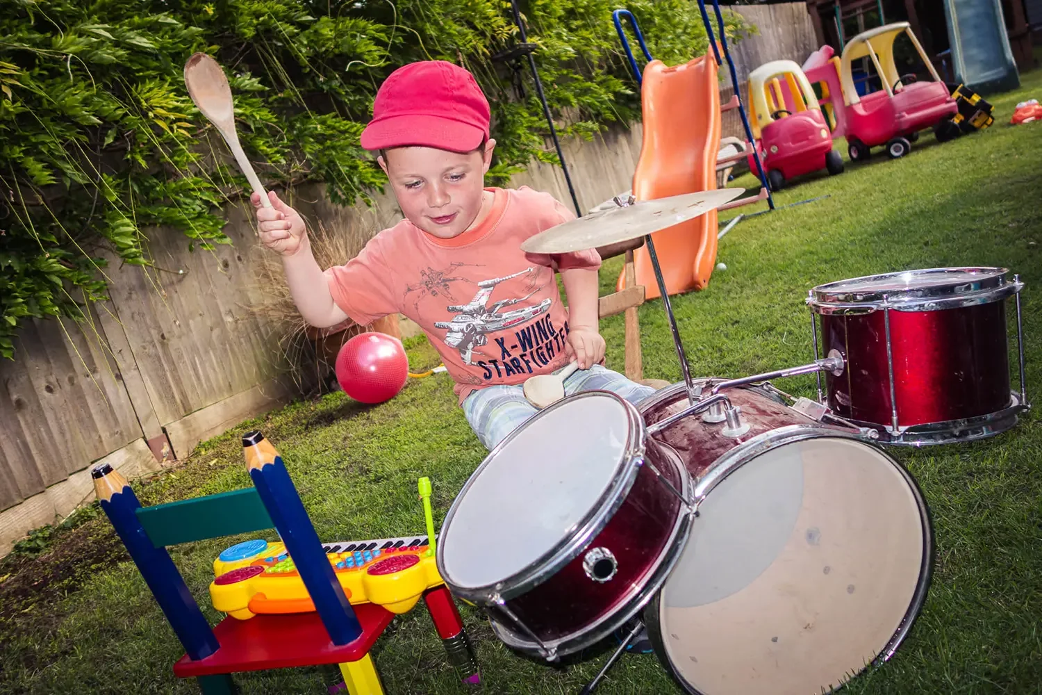 A young boy wearing a red cap and a Starfighter t-shirt plays a drum set outdoors in a backyard, surrounded by toys and a wooden fence with green foliage.