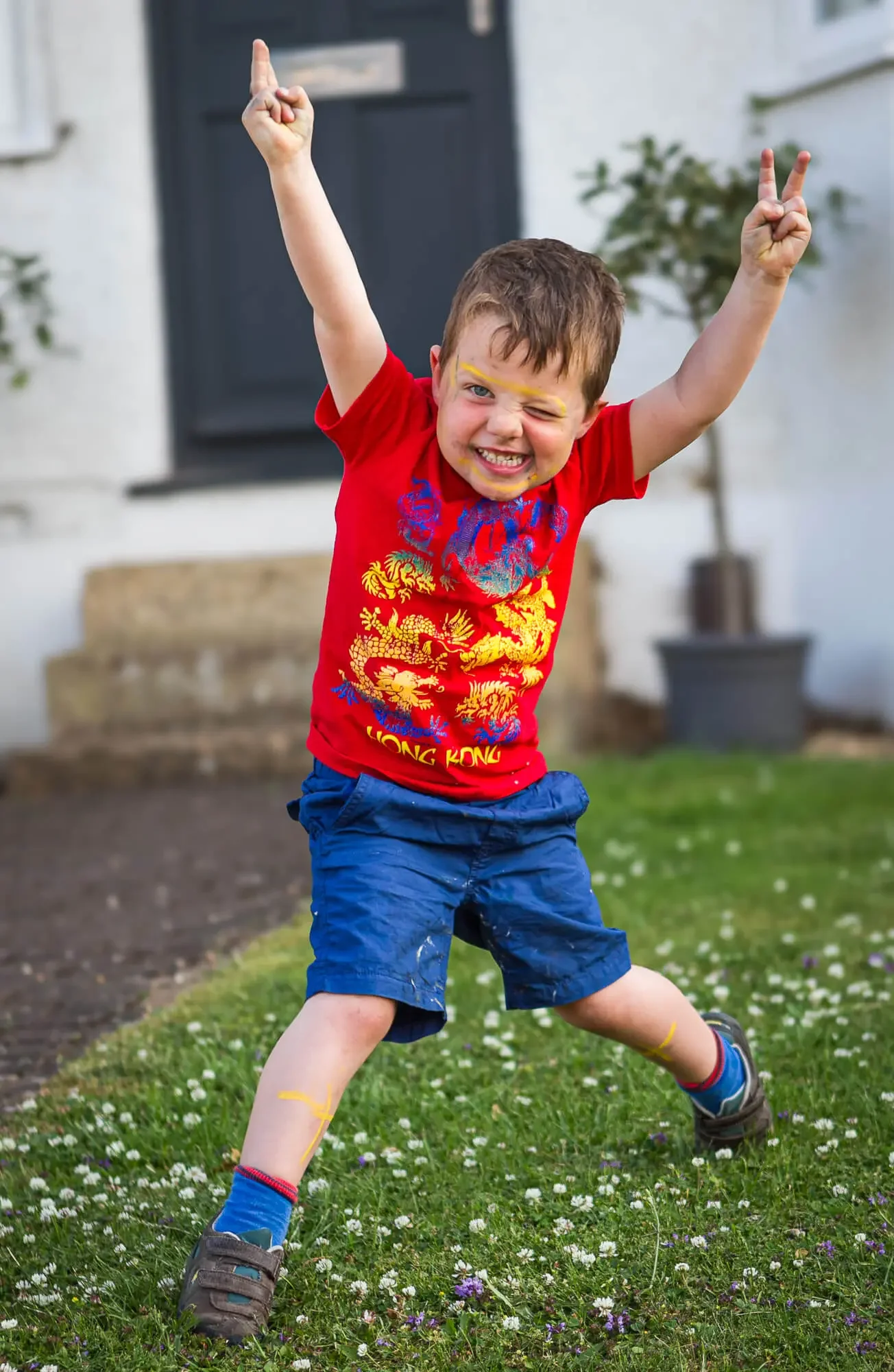 A young boy with face paint and a big smile, standing outdoors on a lawn with small flowers, raising his arms with peace signs, dressed in a red shirt with a dragon design and blue shorts.