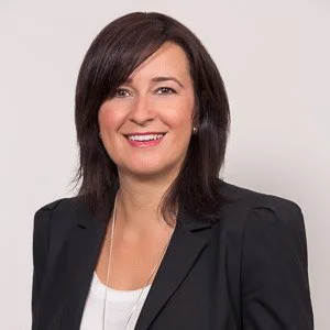 Portrait of a woman with shoulder-length dark brown hair, wearing a black blazer over a white top, smiling against a plain white background.
