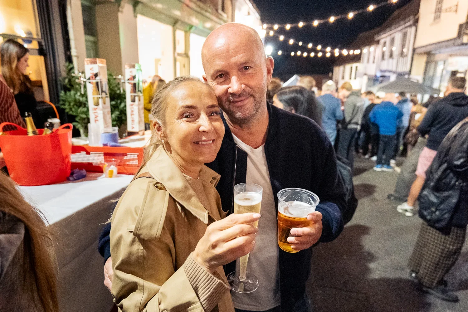 A happy couple pose with drinks in a packing George Street at night under strings of festoon lights, with Tudor buildings on either side