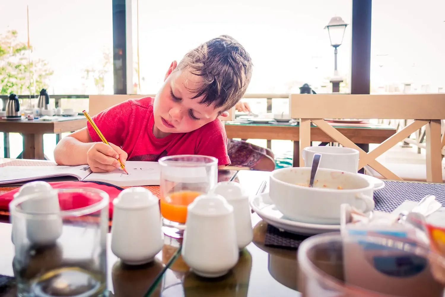 A young boy in a red shirt sitting at a table with breakfast dishes, drawing in a notebook with a yellow pencil.