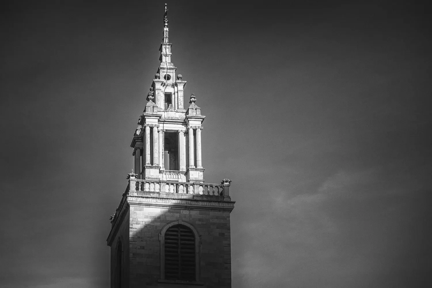 Ornate classical church tower and spire rising against a grey sky in the City of London, strong side light picking out the carved stonework, black and white