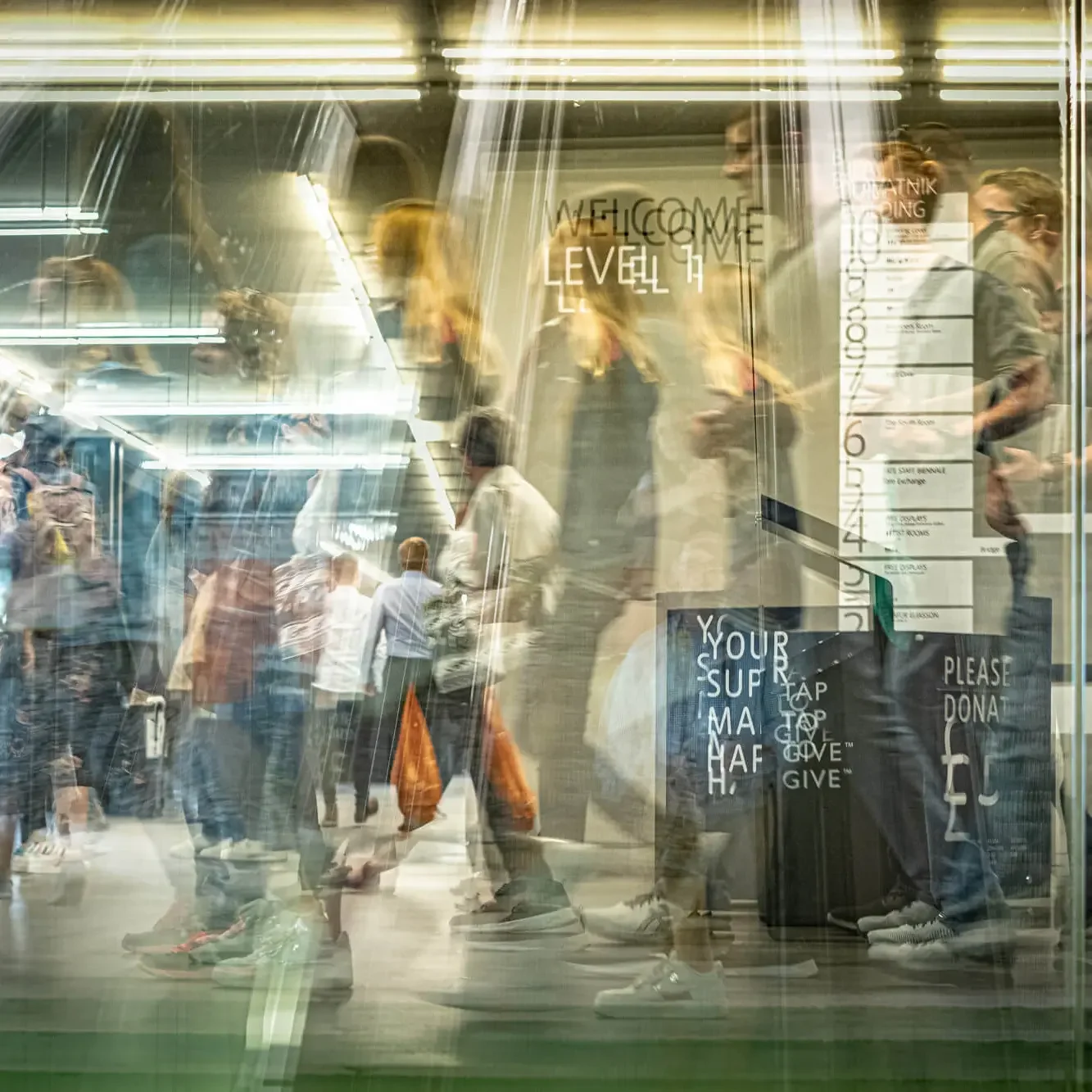 Multiple people walking through an indoor space with reflections on glass, illuminated signage including a welcome sign to level 7, and donation and support signs.
