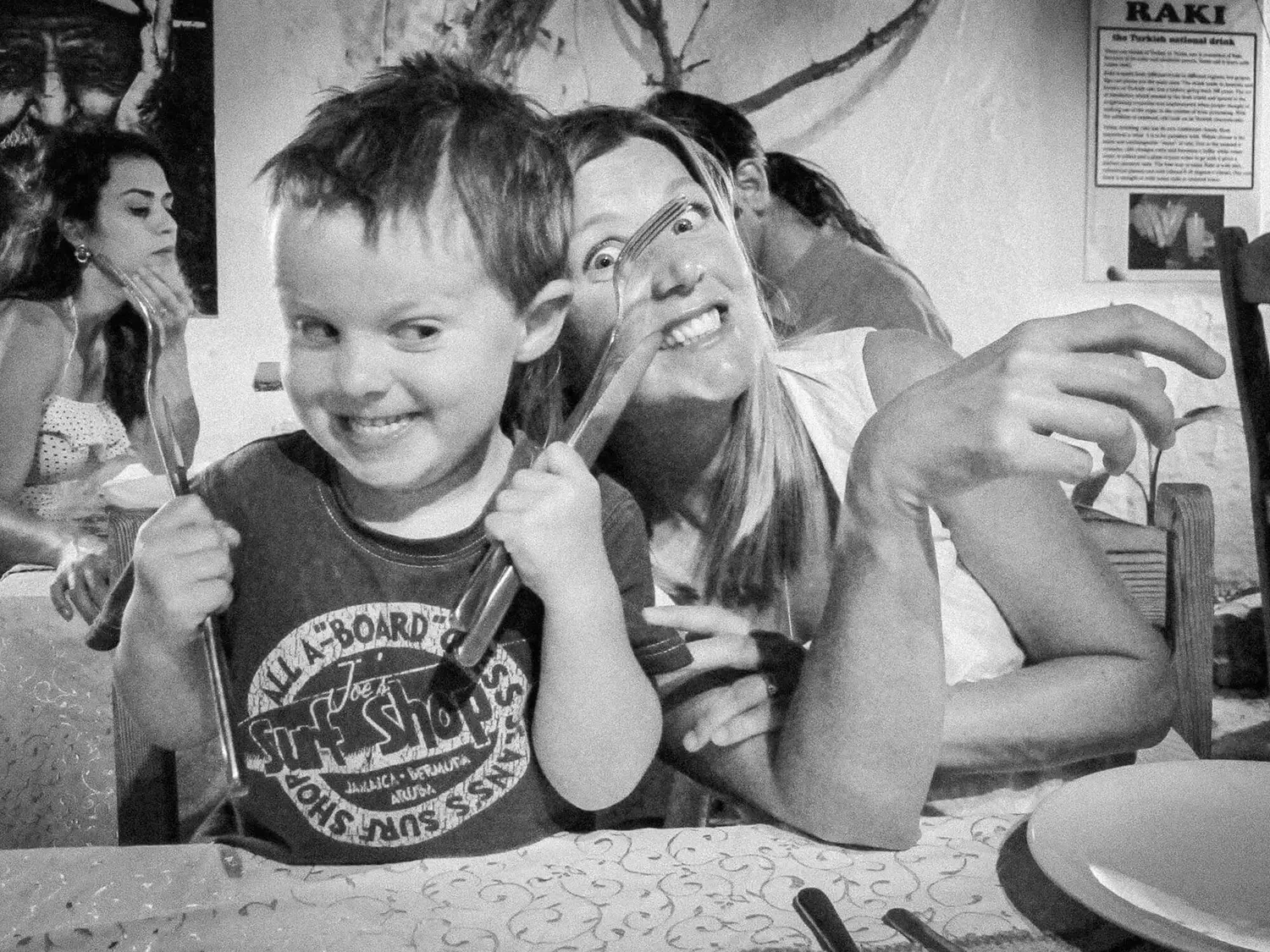 A young boy and woman sitting at a table, smiling and playful, with the boy holding a fork and a knife. Other people are in the background, and there is a sign on the wall with the word 'RAKI'.