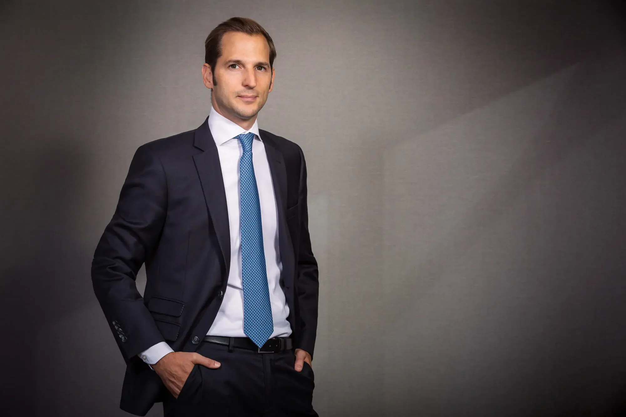 A man in a black suit, white shirt, and blue tie standing confidently with hands in pockets against a neutral gray background.