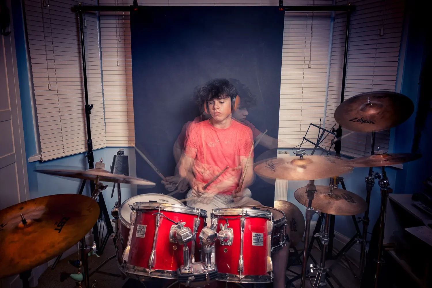 Multiple exposure portrait of a young drummer playing a red Yamaha kit with Zildjian cymbals, wearing orange headphones, with ghosted motion blur conveying rhythm and energy against a dark studio backdrop