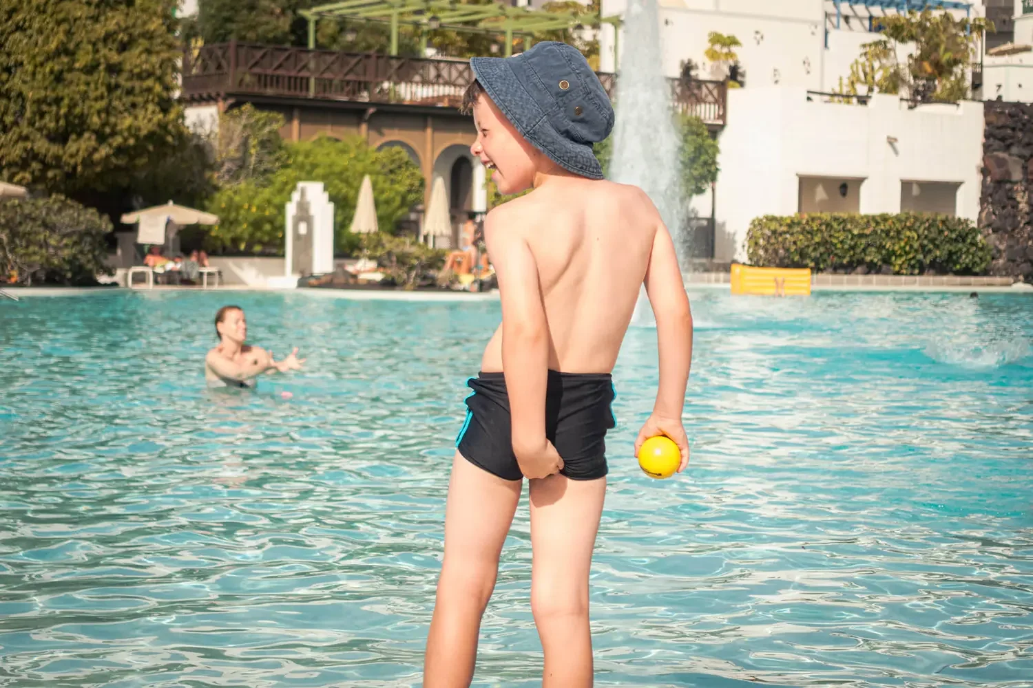 A young boy with a blue hat and black swim shorts standing by a swimming pool, holding a yellow ball behind his back, smiling and looking at a woman playing in the water.