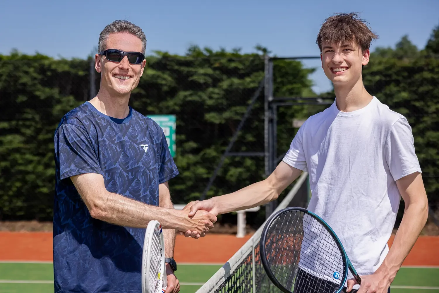 Tennis coach and young male player shaking hands at the net on an outdoor court at Greenwood Park, St Albans, on a sunny day.