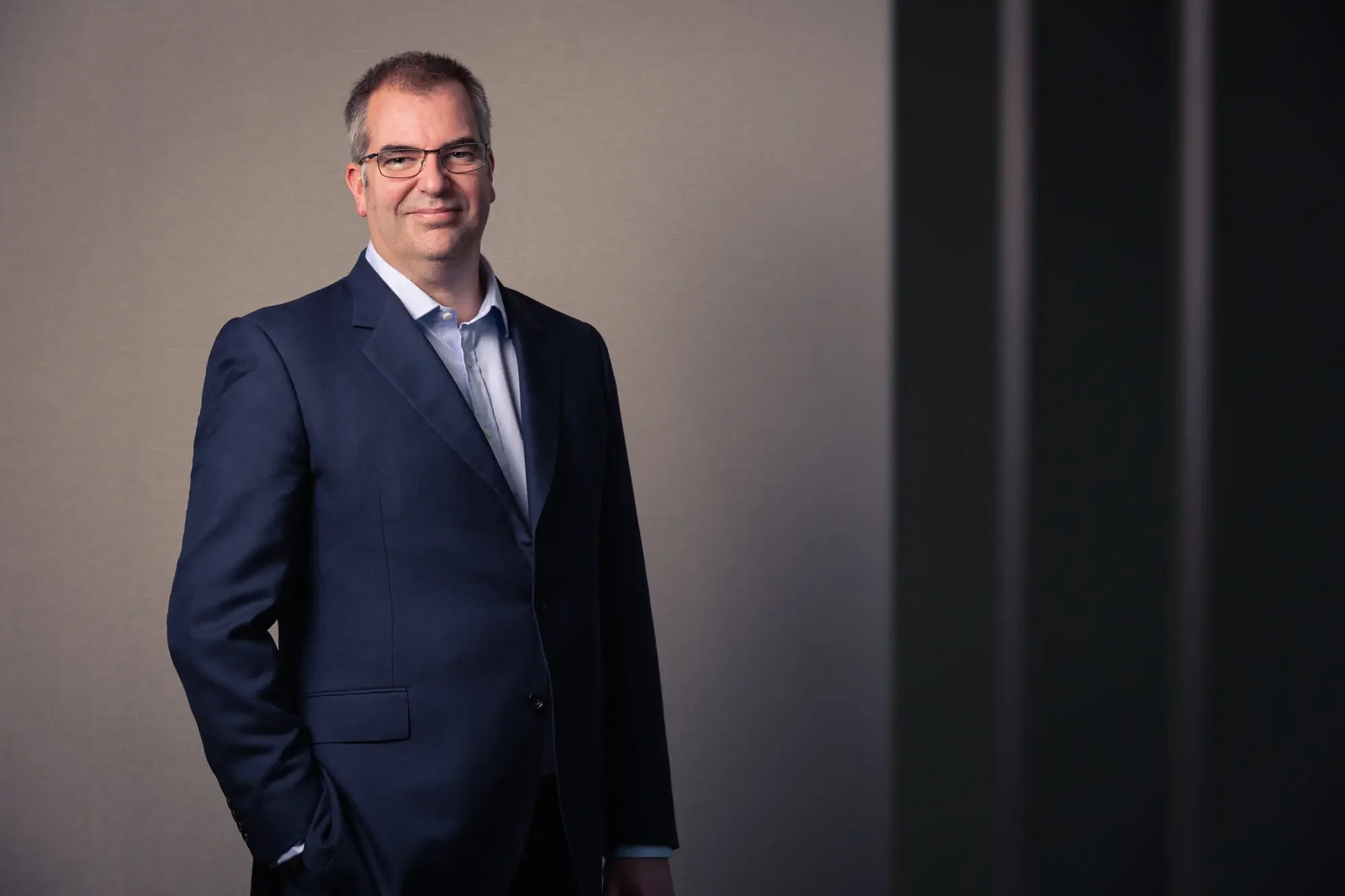 A man in a navy blue suit, white shirt, and glasses standing against a neutral background with a dark object on the right side.