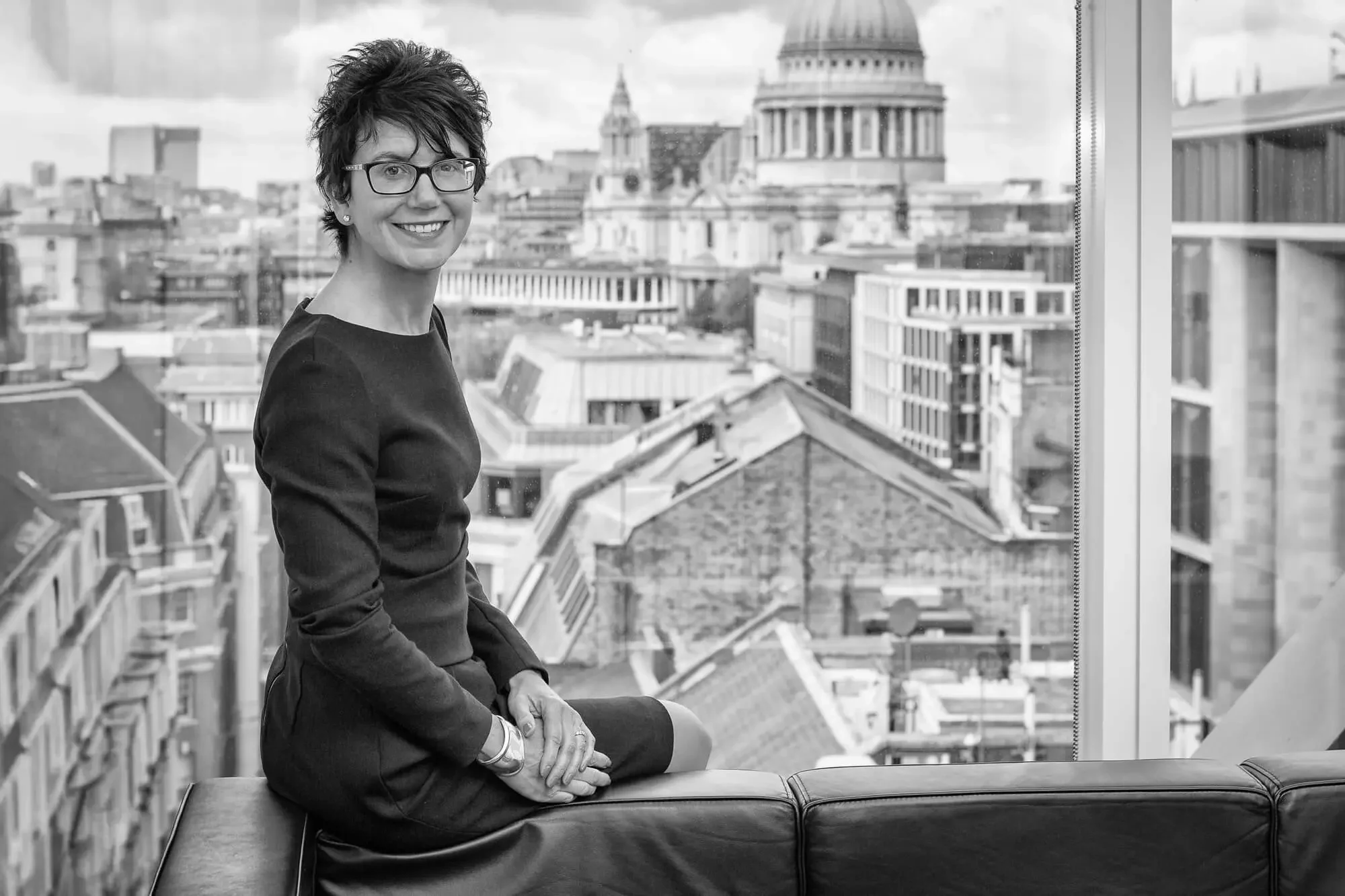 A woman with glasses and a short haircut, smiling while sitting on a bench in front of a large window overlooking a cityscape with historic buildings and a domed government building.