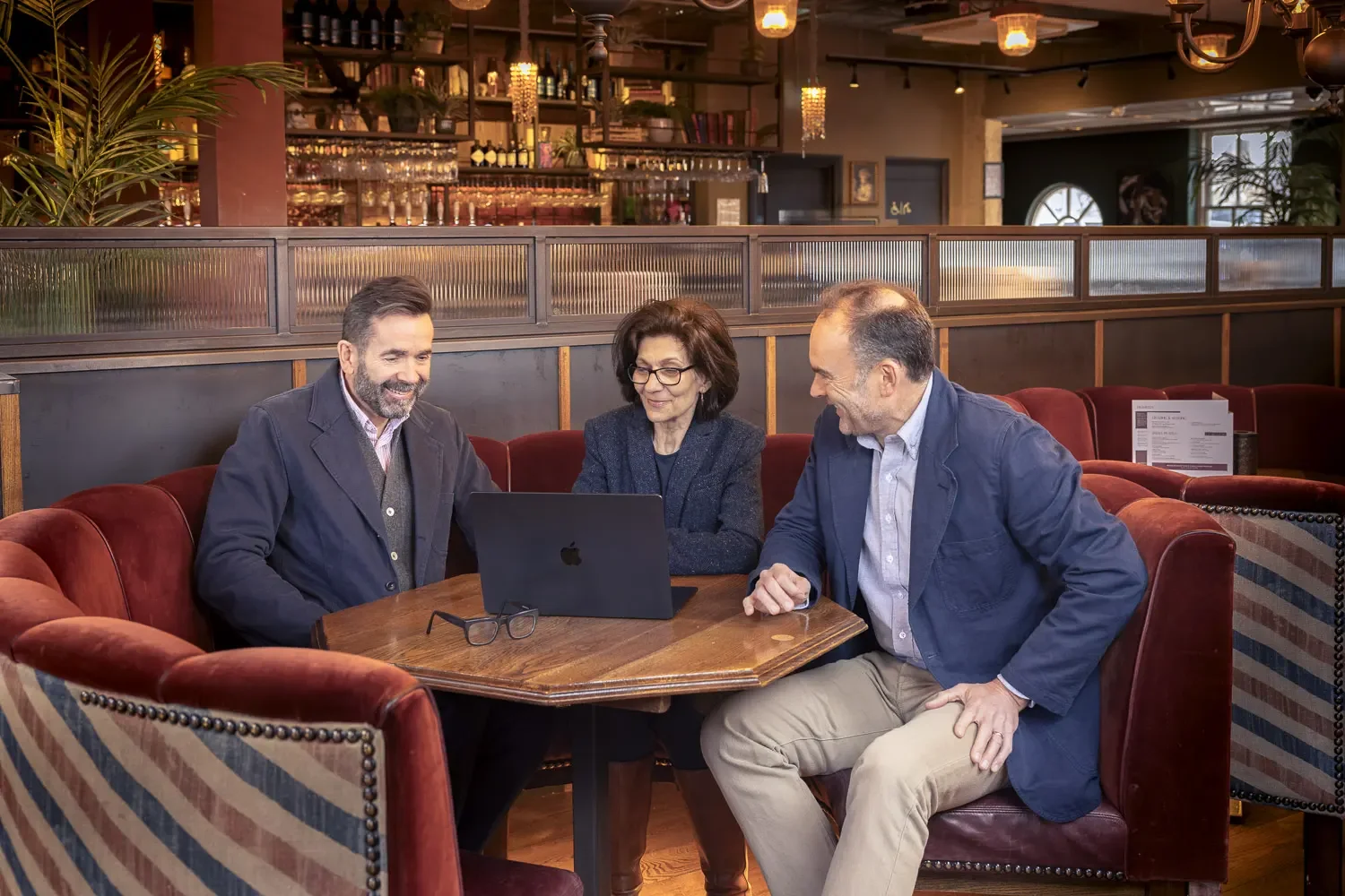 Three middle-aged adults, two men and a woman, sitting at a wooden table in a restaurant or cafe, looking at a laptop and smiling, engaged in a discussion.