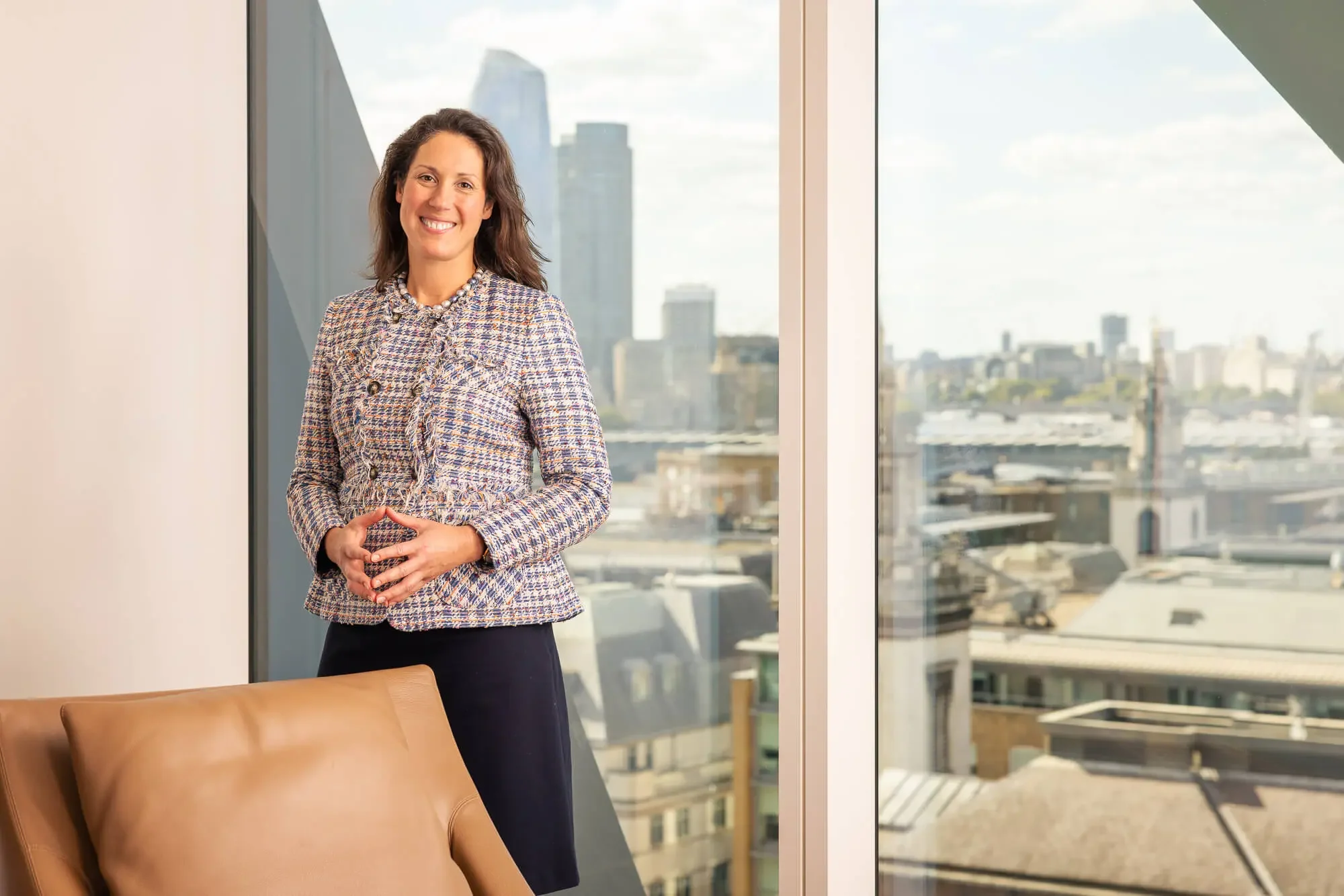 A woman with shoulder-length brown hair stands by a large window with a city skyline in the background, smiling and wearing a patterned blazer and a dark skirt.