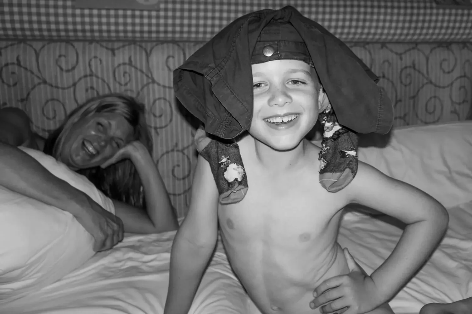 A young boy with a large hat and socks smiling confidently, sitting shirtless on a bed. In the background, a woman is lying on the bed, resting her head on her hand and smiling.