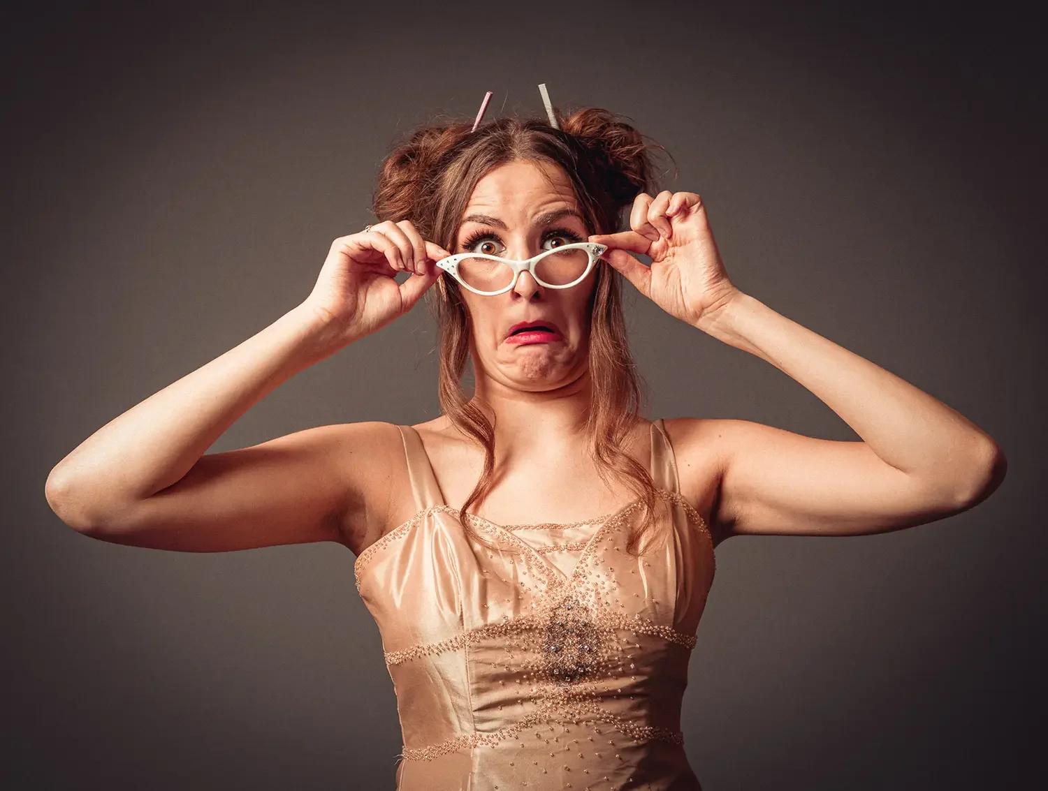 A woman with messy hair and a surprised expression, wearing a spaghetti strap dress and holding eyeglasses, with her mouth turned down in a humorous pout. Ready for panto at St Albans, Abbey Theatre.