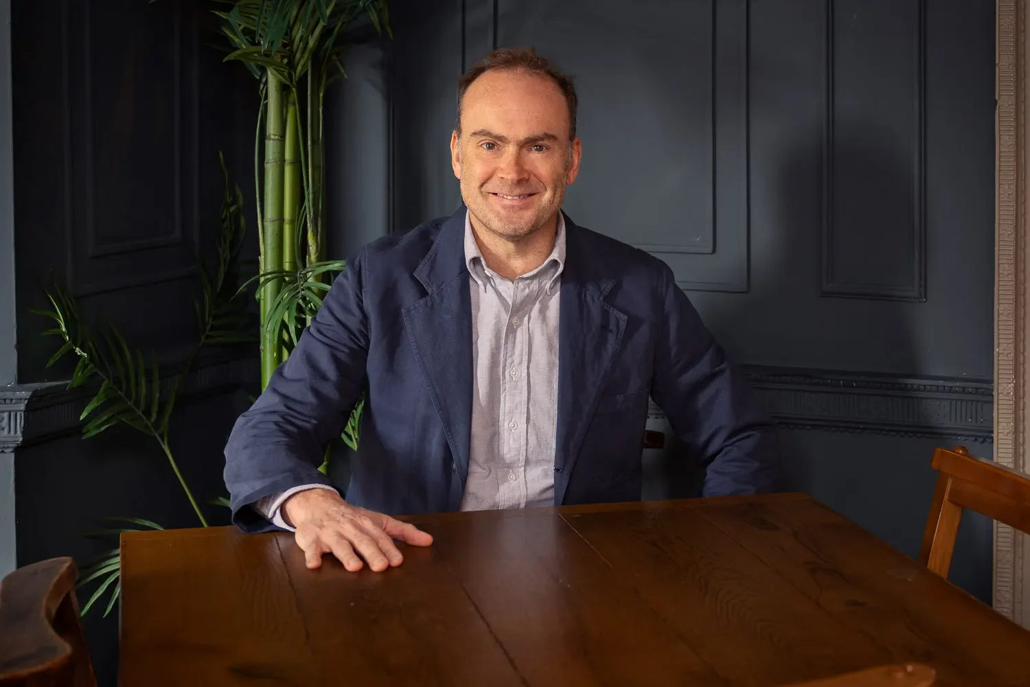 Male business professional in a navy blazer seated at a wooden table in a blue panelled room — personal branding headshot, St Albans